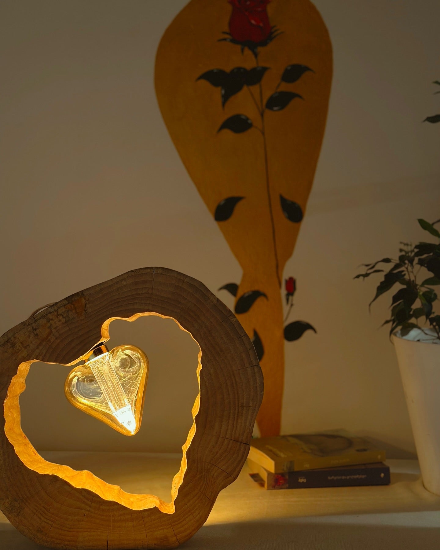 A table lamp with a heart-shaped design, made from wood and illuminated from the inside, casting a warm glow. The lamp is placed in a room with a decorative wall and a potted plant.