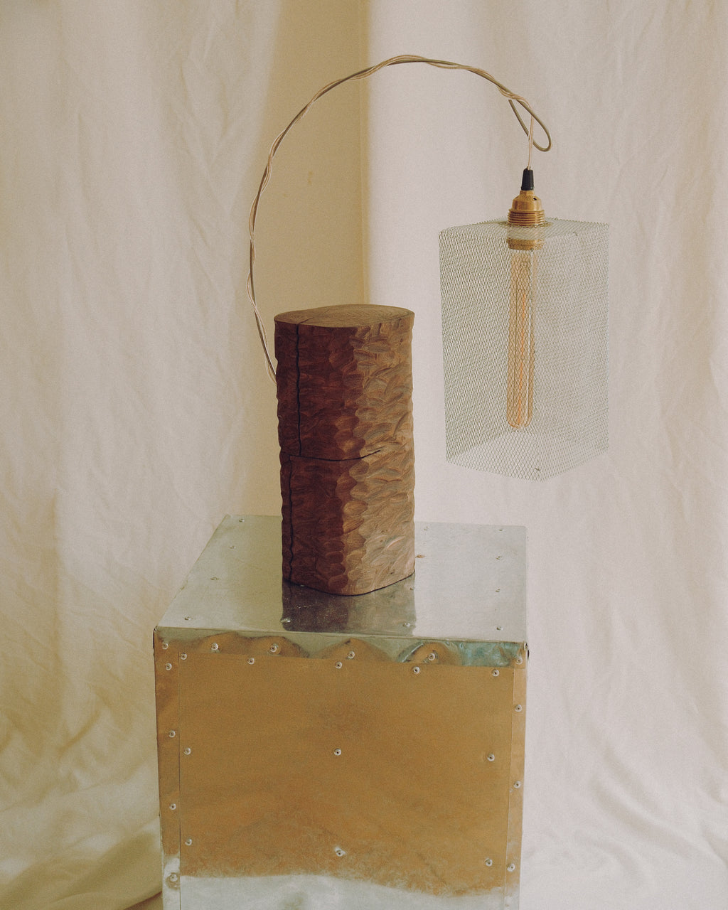 A rustic industrial-style pendant light with a hammered brown metal cylindrical shade, a clear glass rectangular pendant box, and a thin metal hanging loop, displayed on a reflective metallic pedestal against a cream fabric backdrop.