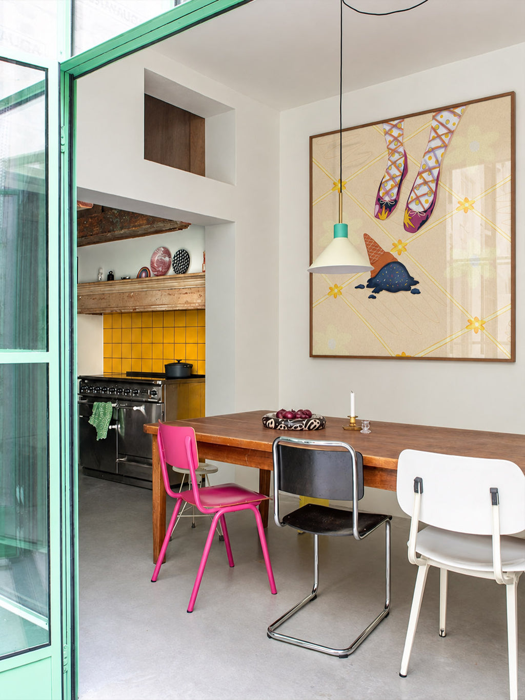 A dining area with a wooden table and three chairs (pink, black-and-silver, and white) in a modern room, with a large framed art print on the wall depicting patterned socks by DAE