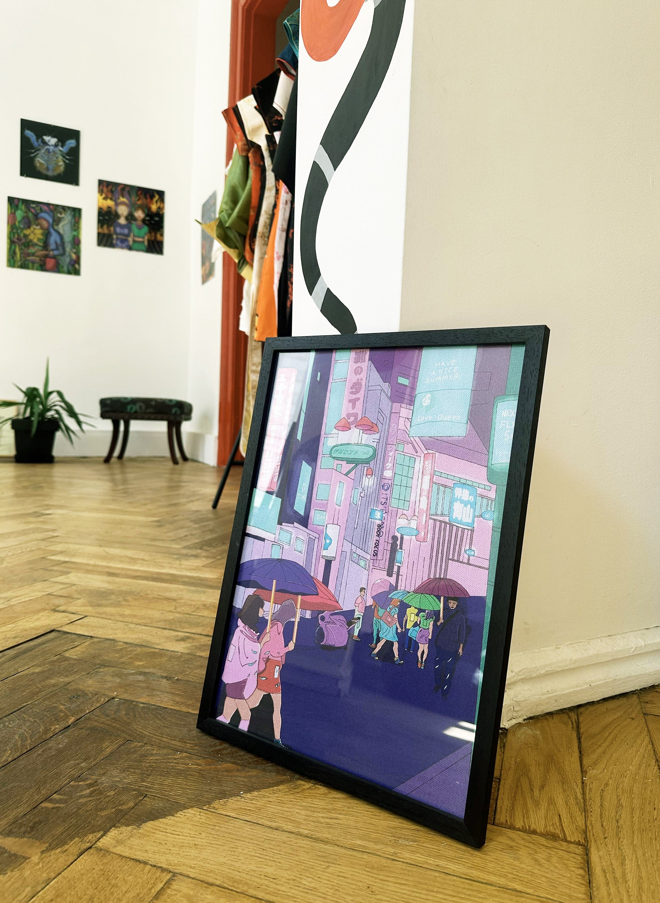 A poster featuring an illustration of a cityscape with a prominent purple building, displayed in an art gallery setting with various other artworks in the background. Poster “Japan” by Lasha Lezhava – Georgian art print blending Tbilisi’s rhythm and Japanese serenity, velvet version by DAE • დაე.