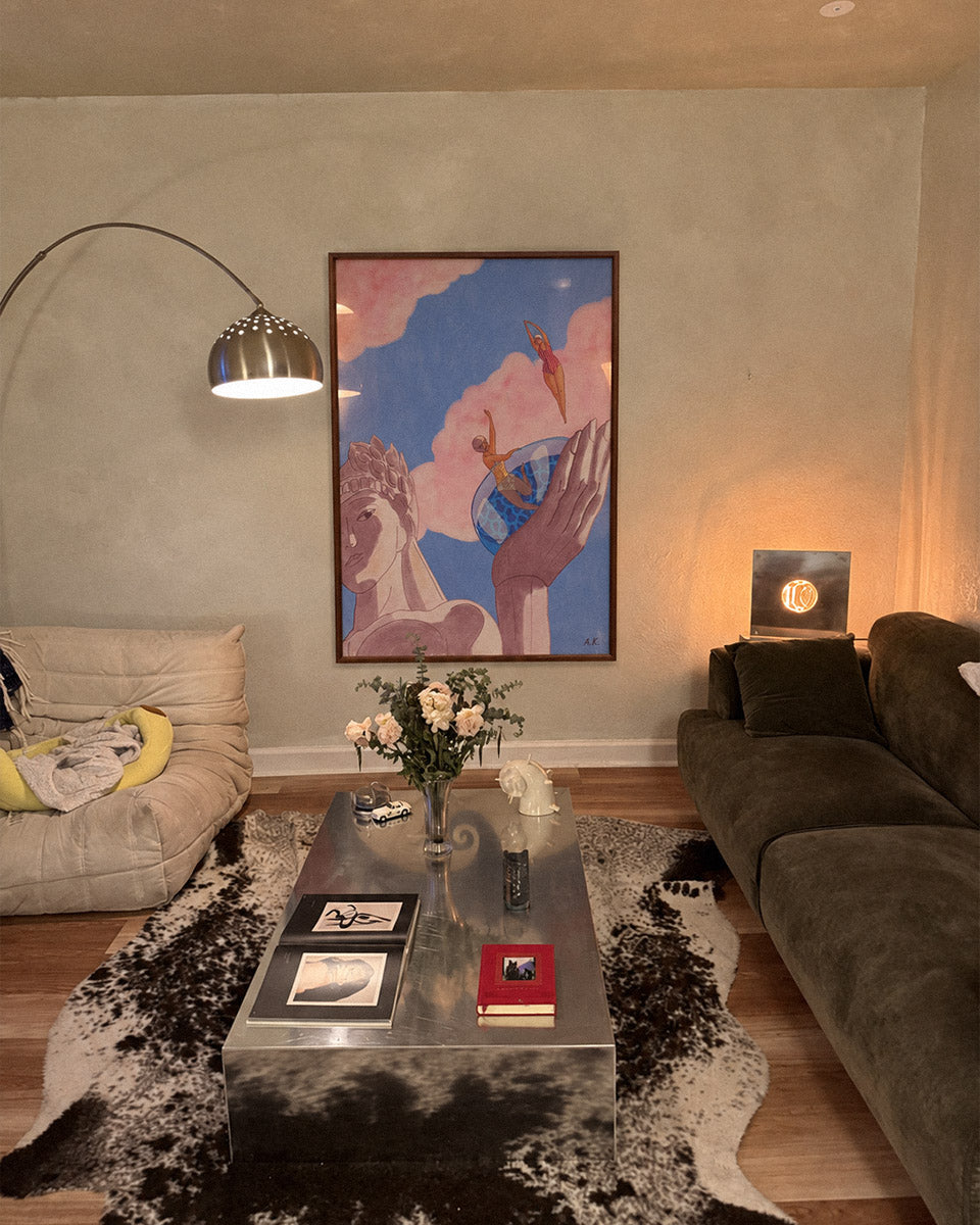 A framed art print featuring a surreal painting of a hand holding a winged figure under a blue sky with pink clouds, mother of georgia poster by DAEdisplayed on a beige wall in a modern living room setting.
