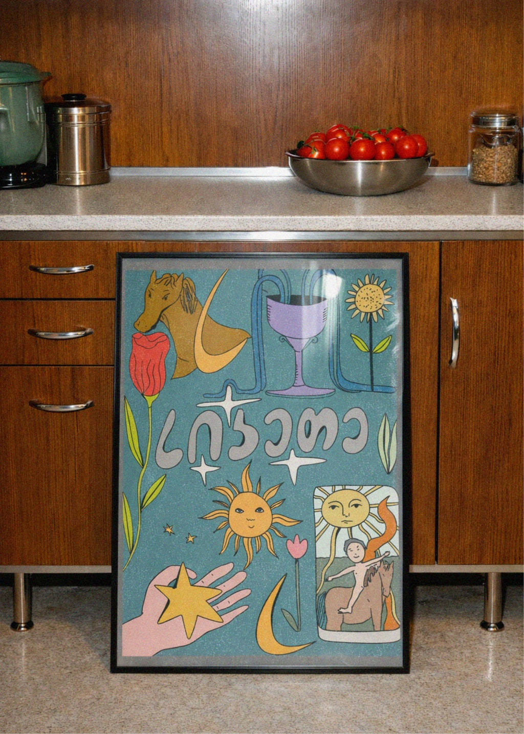 Decorative framed artwork on a kitchen counter with wooden cabinets and a bowl of tomatoes in the background.