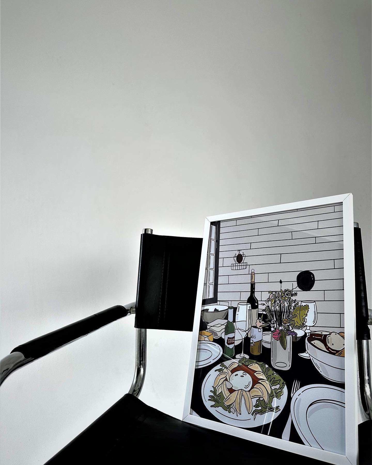 Framed artwork of a dining scene with a chair in front of it. Table art poster by Oliko Tkebuchava - minimalist wall decor.