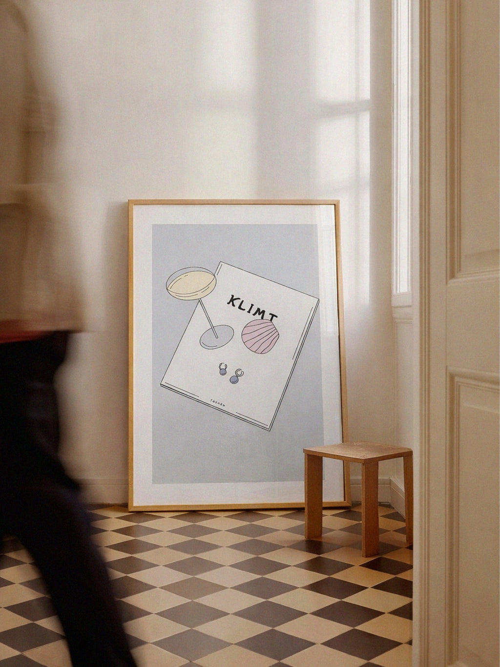 Framed artwork on a wall with a checkered floor and wooden stool.