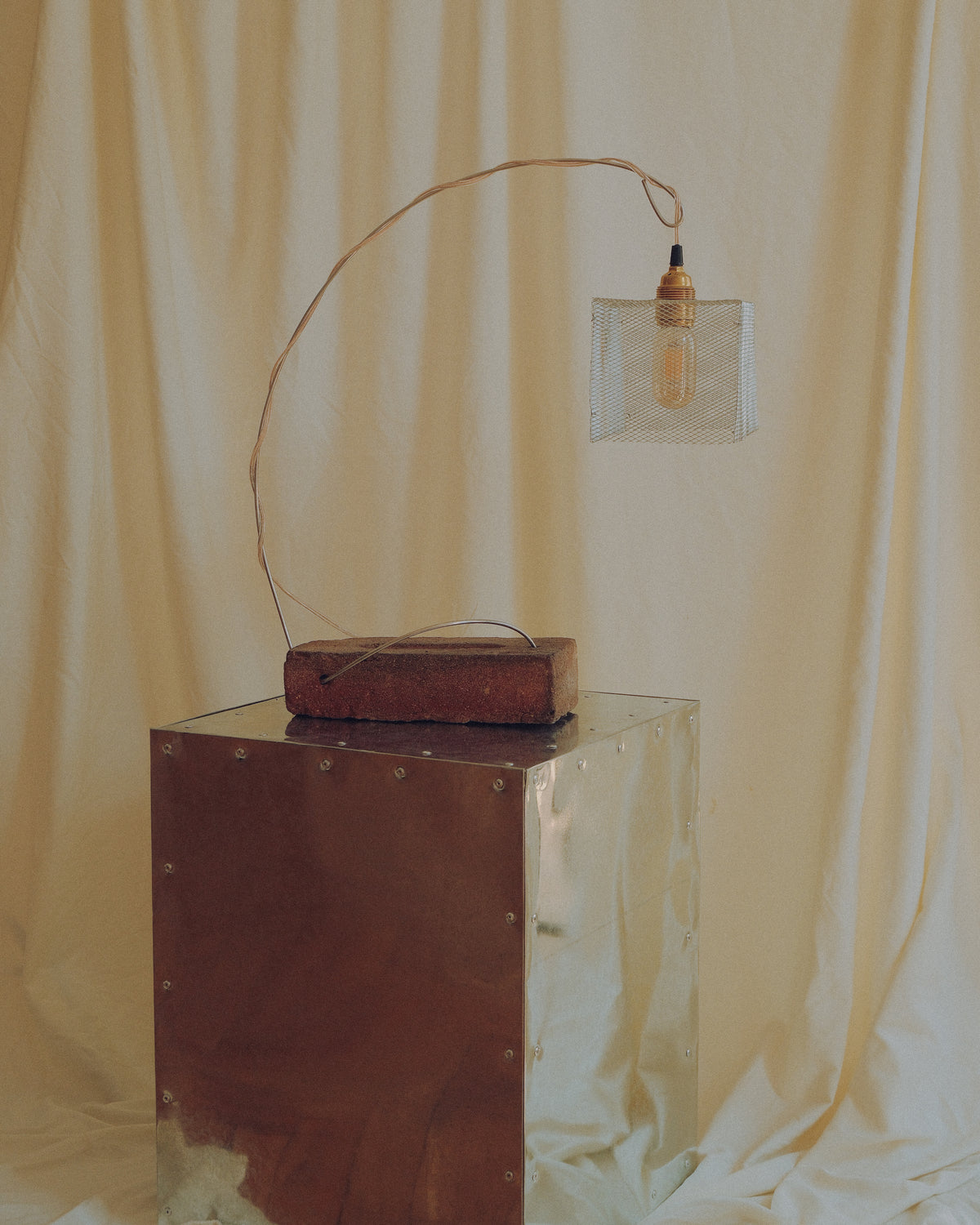 A rustic industrial-style table lamp with a curved metal arm, a wooden base, and a clear glass shade, displayed on a metallic pedestal against a draped cream fabric backdrop.