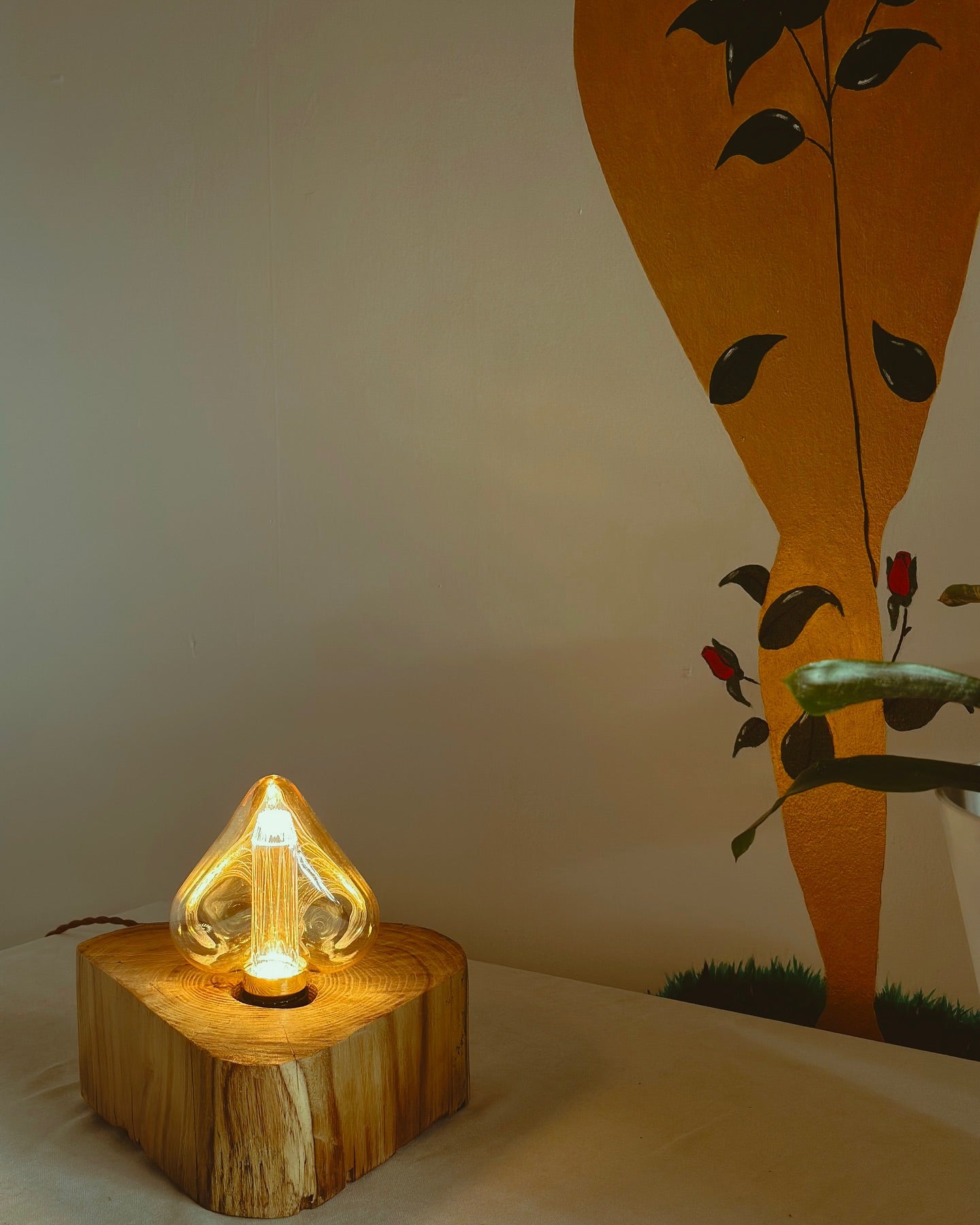 A handmade lamp with a unique design, featuring a wooden base and an illuminated, flower-shaped lampshade, placed on a table with a wall painting in the background.