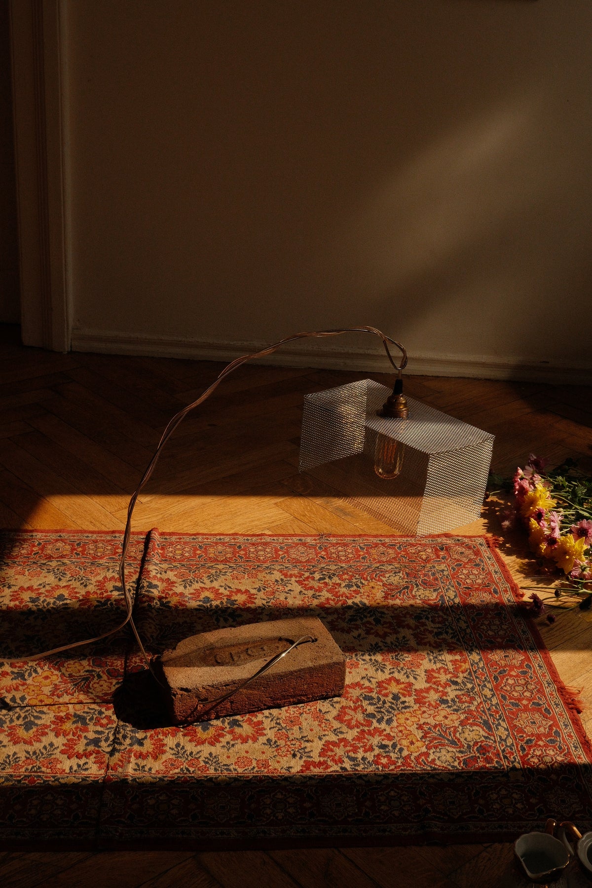 A rustic, industrial-style pendant light with a long, curved wire frame and a clear, geometric acrylic block shade, resting on a patterned rug in a dimly lit room.