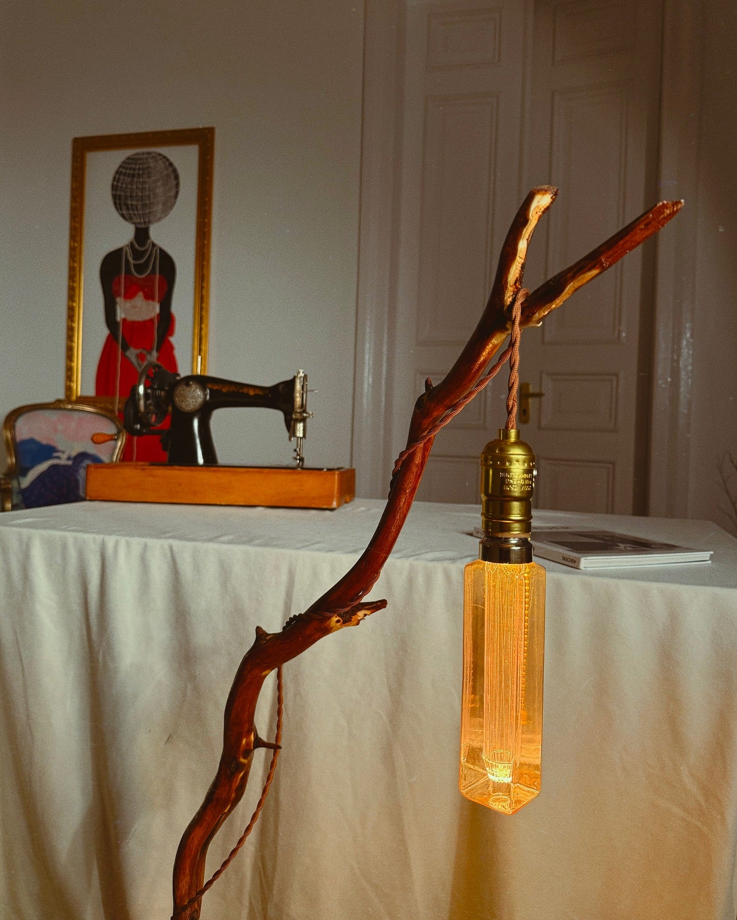 A handmade wooden lamp with a branch-like structure and a glowing bulb, displayed in a room setting with a table and chair in the background.