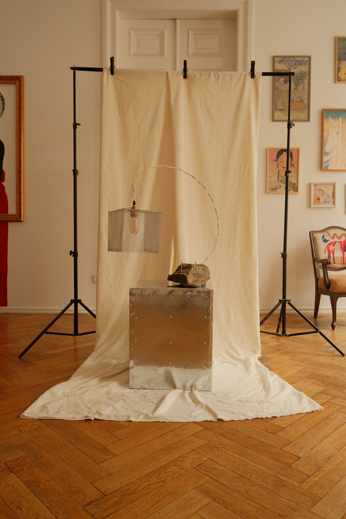 A sculptural lamp with a square metal shade and a curved metal arm is displayed on top of a metallic cube pedestal in an indoor studio setup. A draped cream fabric backdrop is suspended between two black lighting stands on a wood floor, with framed wall art and a chair visible in the room.