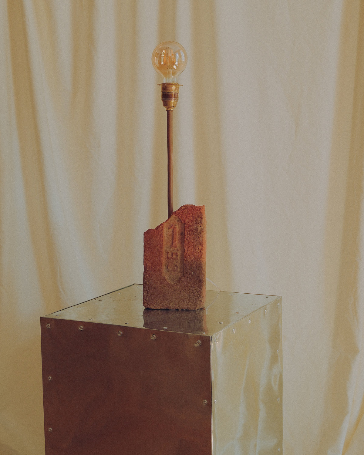A vintage-style table lamp with a clear glass bulb on a long dark stem, mounted on a rusted metal block that sits atop a reflective metal pedestal. The backdrop is a draped cream fabric.