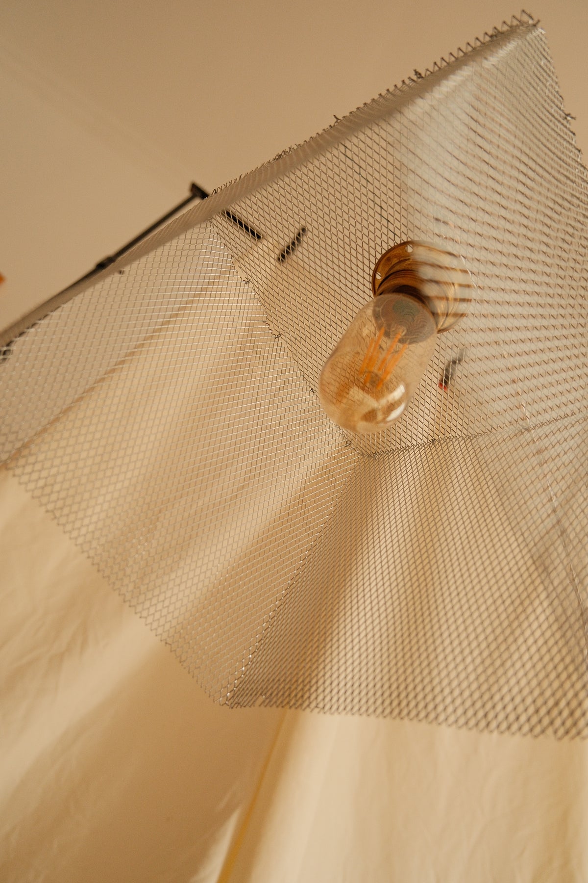A close-up of a textured, metallic mesh fabric panel with a warm-toned Edison bulb hanging in the center, set against a draped beige fabric backdrop.
