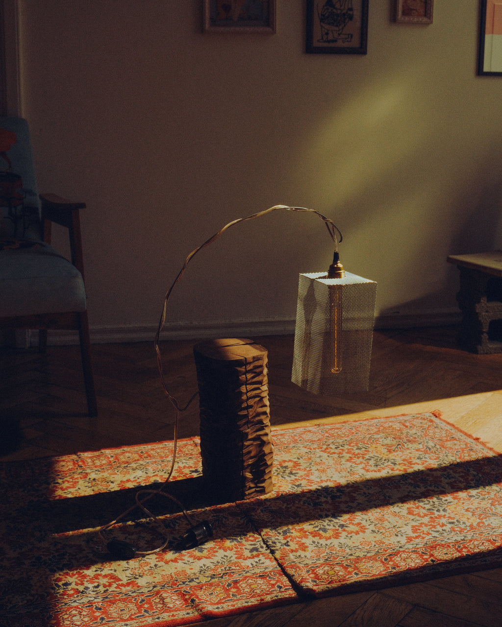 A modern sculptural floor lamp with a tall rectangular white shade, a curved copper-toned wire frame, and a stacked wood base, set in a dimly lit room with warm sunlight and shadows.