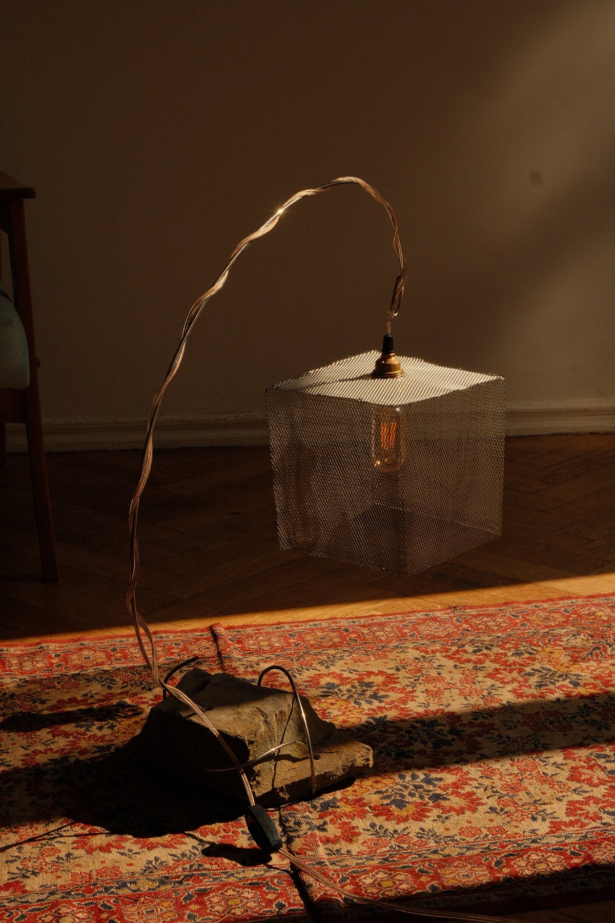 A rustic industrial-style pendant lamp with a square white metal shade, a long curved metal wire frame, and a visible Edison bulb, resting on a patterned red and blue rug in a dimly lit room.
