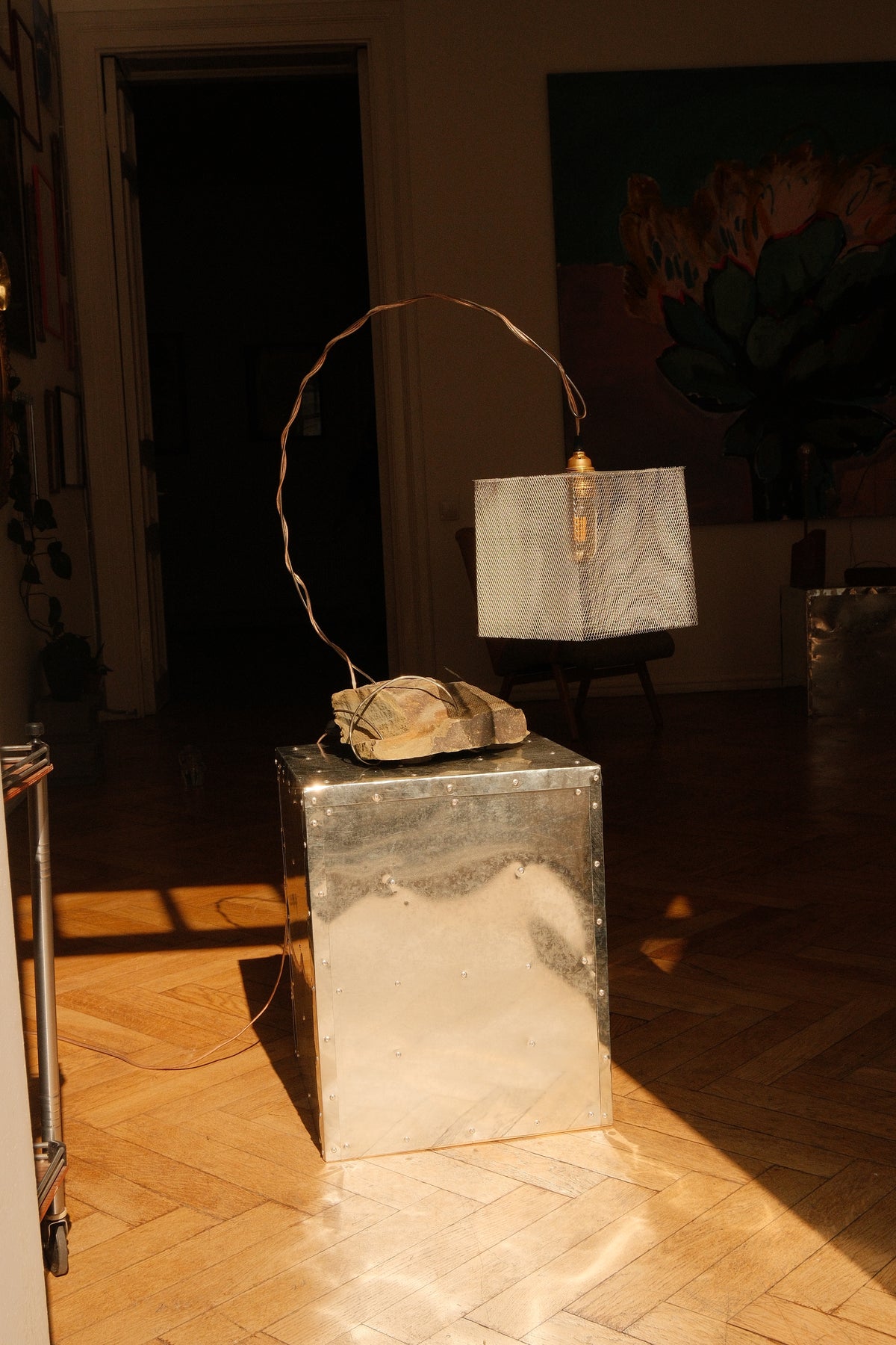 A sculptural table lamp with a rectangular white metal shade, a curved copper wire arm, and a rough stone base, displayed on a metallic pedestal in a sunlit room with a parquet floor.