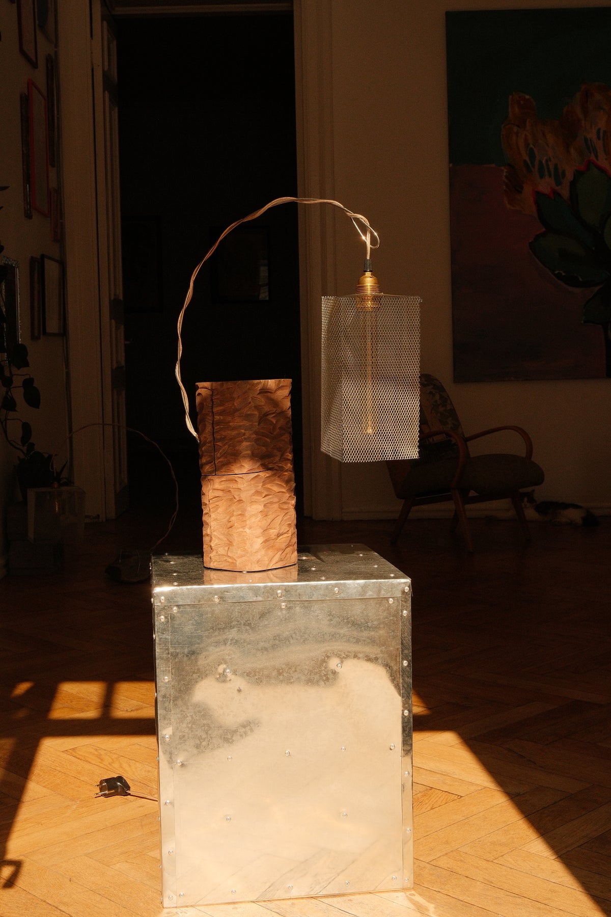 A modern sculptural table lamp featuring a textured brown wood block and a translucent clear glass cylinder, suspended by a thin gold-toned wire, displayed on a metallic silver pedestal in a sunlit room.