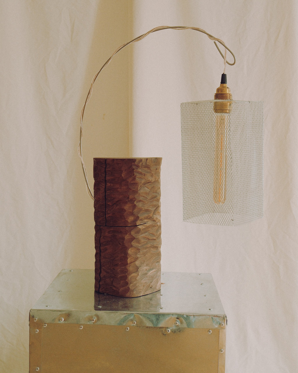 A rustic industrial-style pendant light with a hammered copper-toned cylindrical shade and a hanging brass wire canopy, displayed next to a matching textured metal trunk on a draped beige fabric background.