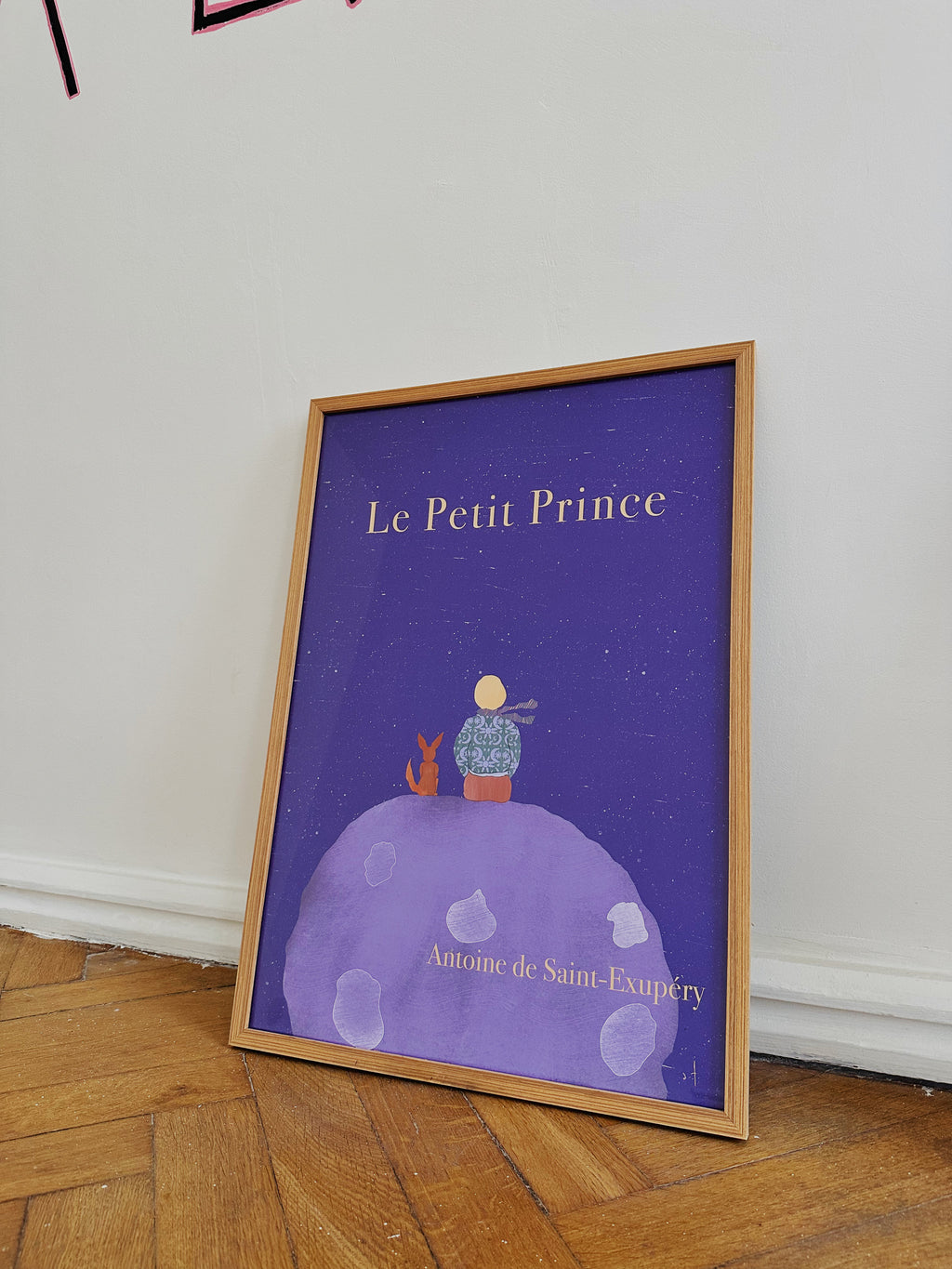 A framed poster of 'Le Petit Prince' by DAE with an illustration of a character sitting on a planet, titled 'Le Petit Prince' at the top leaning against a wall on a wooden floor.