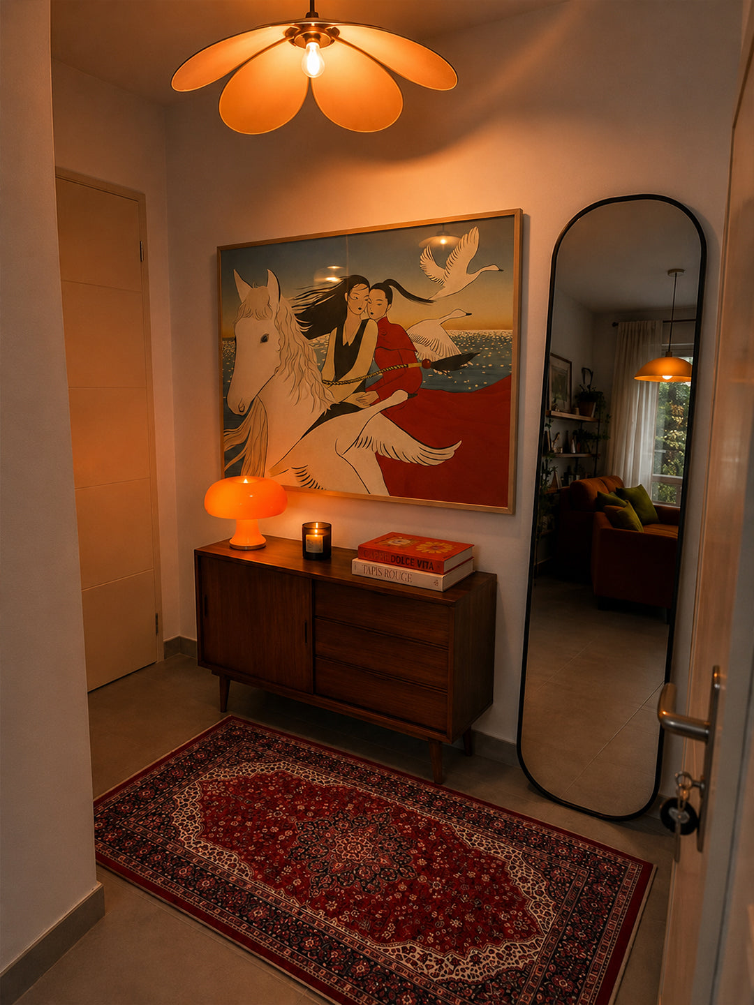 A warm, cozy interior hallway featuring a large framed art print of a woman on a white horse, displayed above a wooden sideboard. The scene is illuminated by a distinctive flower-shaped ceiling light and an orange table lamp, with a red patterned rug on the floor and a tall black-framed mirror on the right.