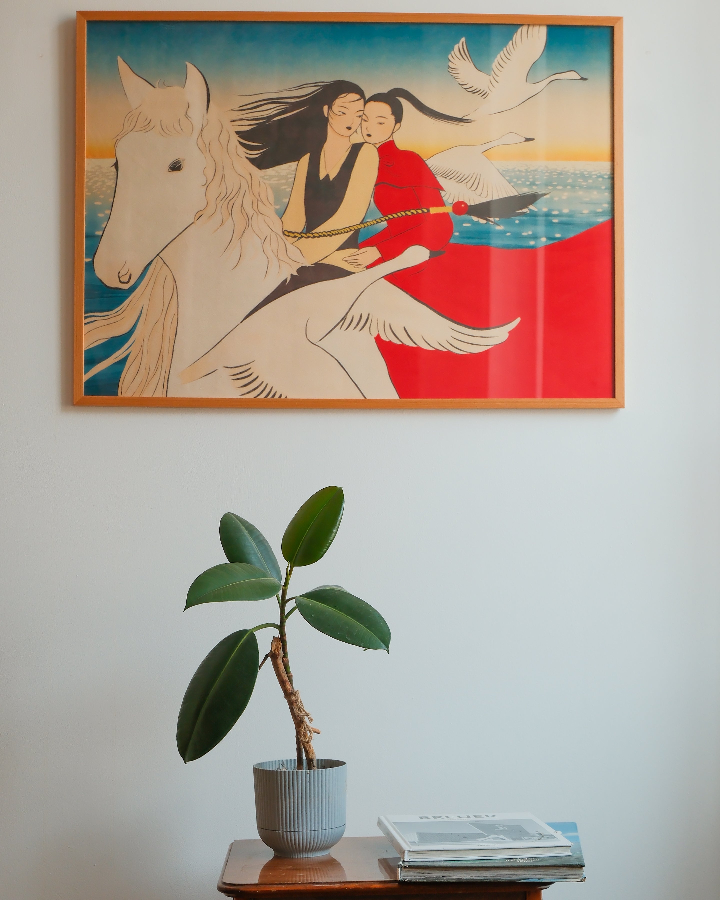 A framed art print depicting a woman riding a white winged horse with birds and a red landscape, hanging on a white wall above a potted plant and books.