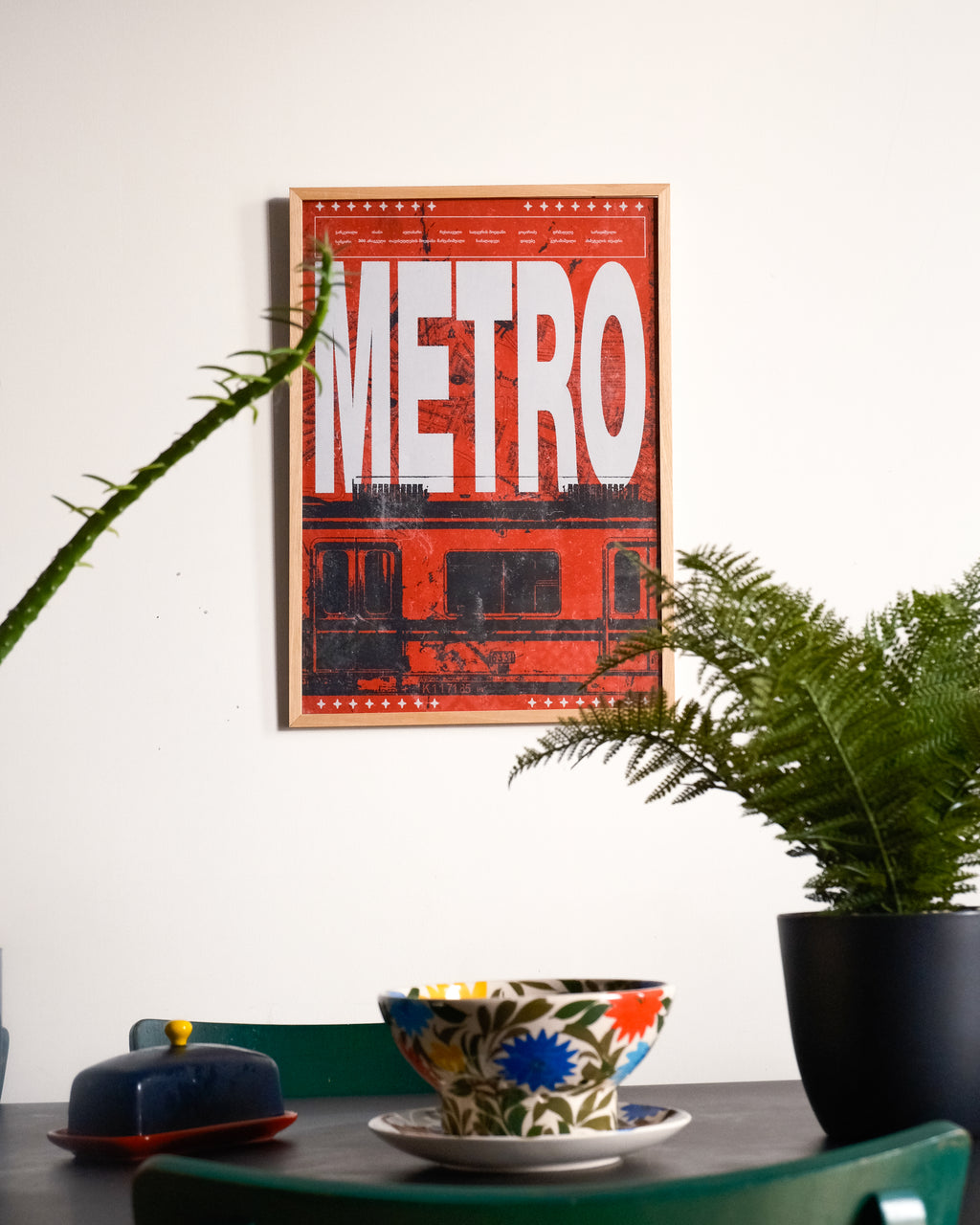Framed poster with 'METRO' text on a wall above a table with a bowl and plant.