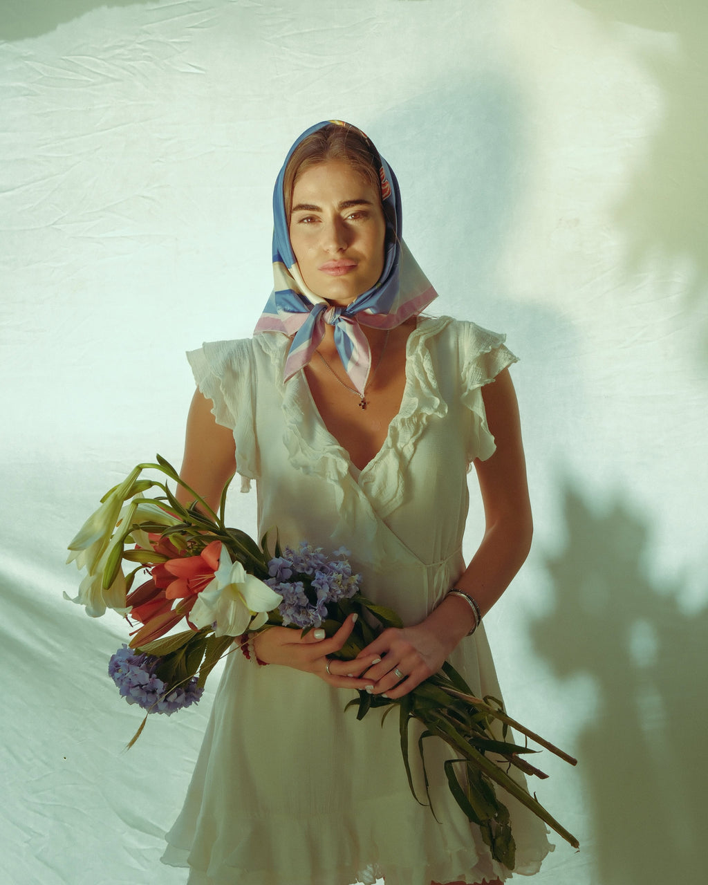 A woman wearing a white ruffled dress and a blue, pink, and white patterned headscarf holds a bouquet of white, red, and purple flowers against a light background with soft shadows.