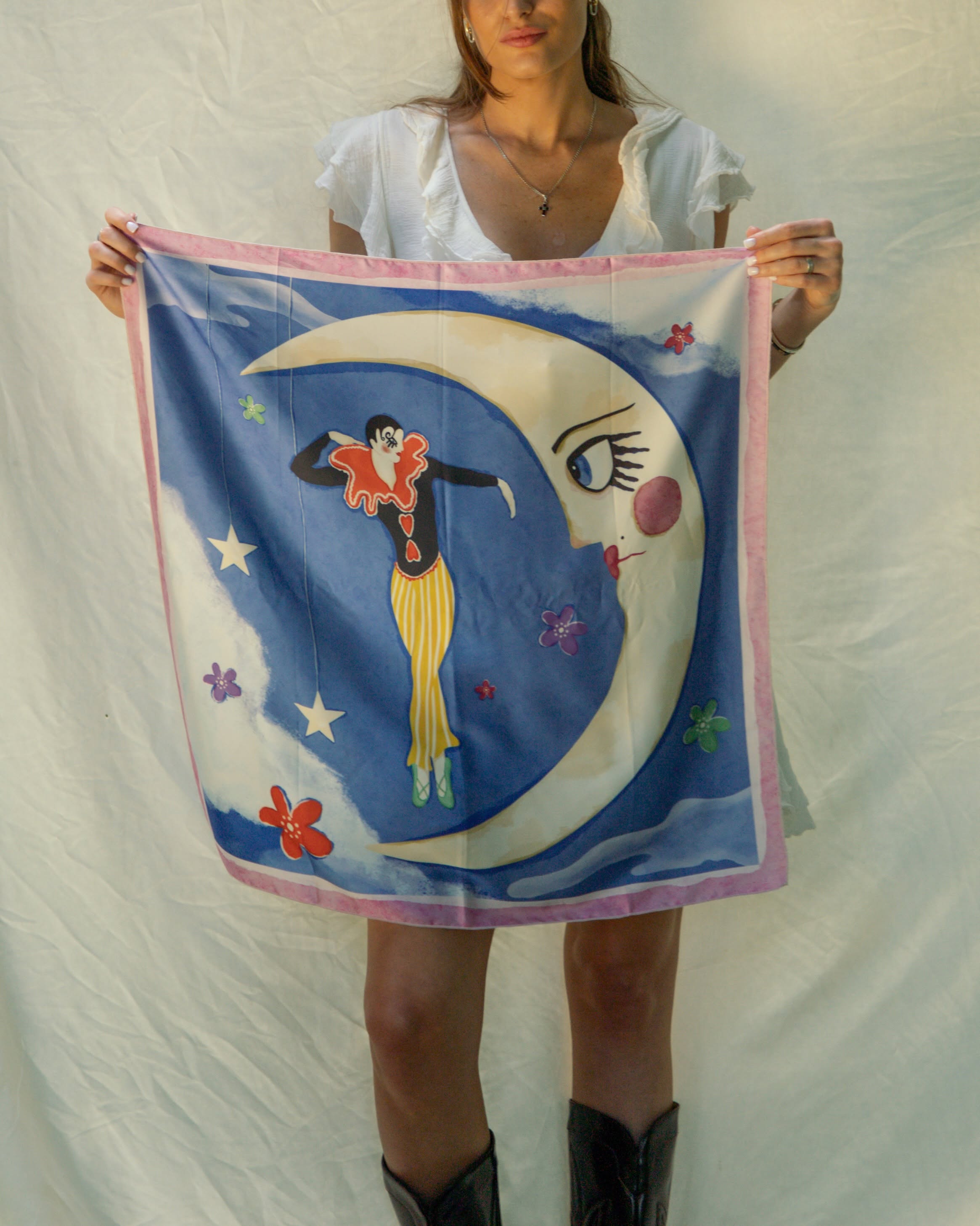 A woman holds up a square scarf featuring a whimsical illustration of a crescent moon face, stars, flowers, and a figure in a red and yellow outfit on a blue background, with a pink border.