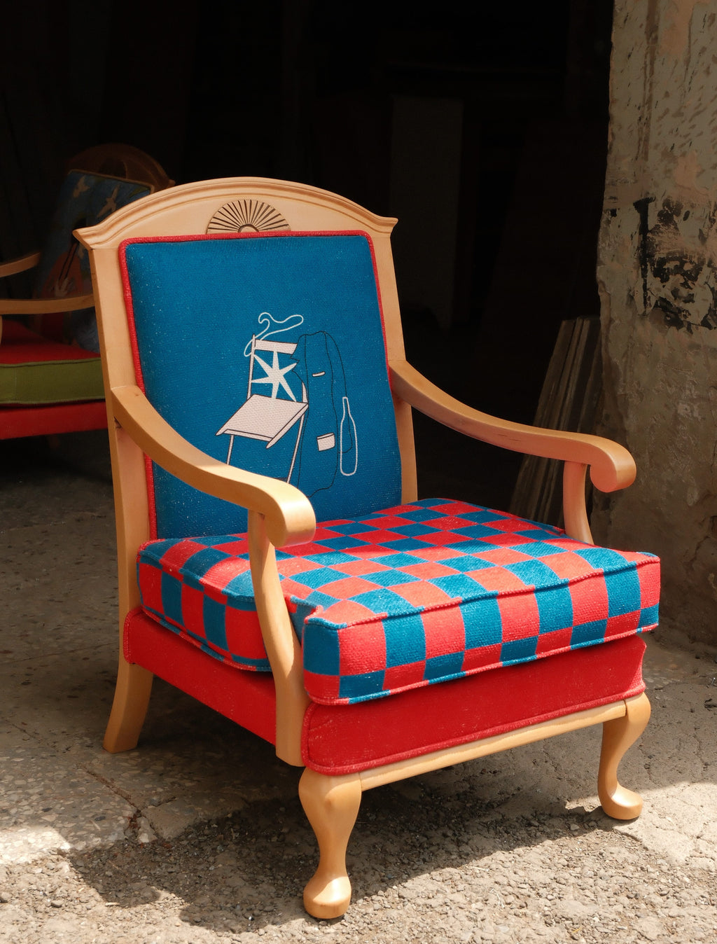 A vintage wooden armchair with a colorful patterned upholstery, featuring a red and blue checkered seat cushion, a solid red upholstered base, and a blue backrest with a white line-art illustration of a smoking figure.