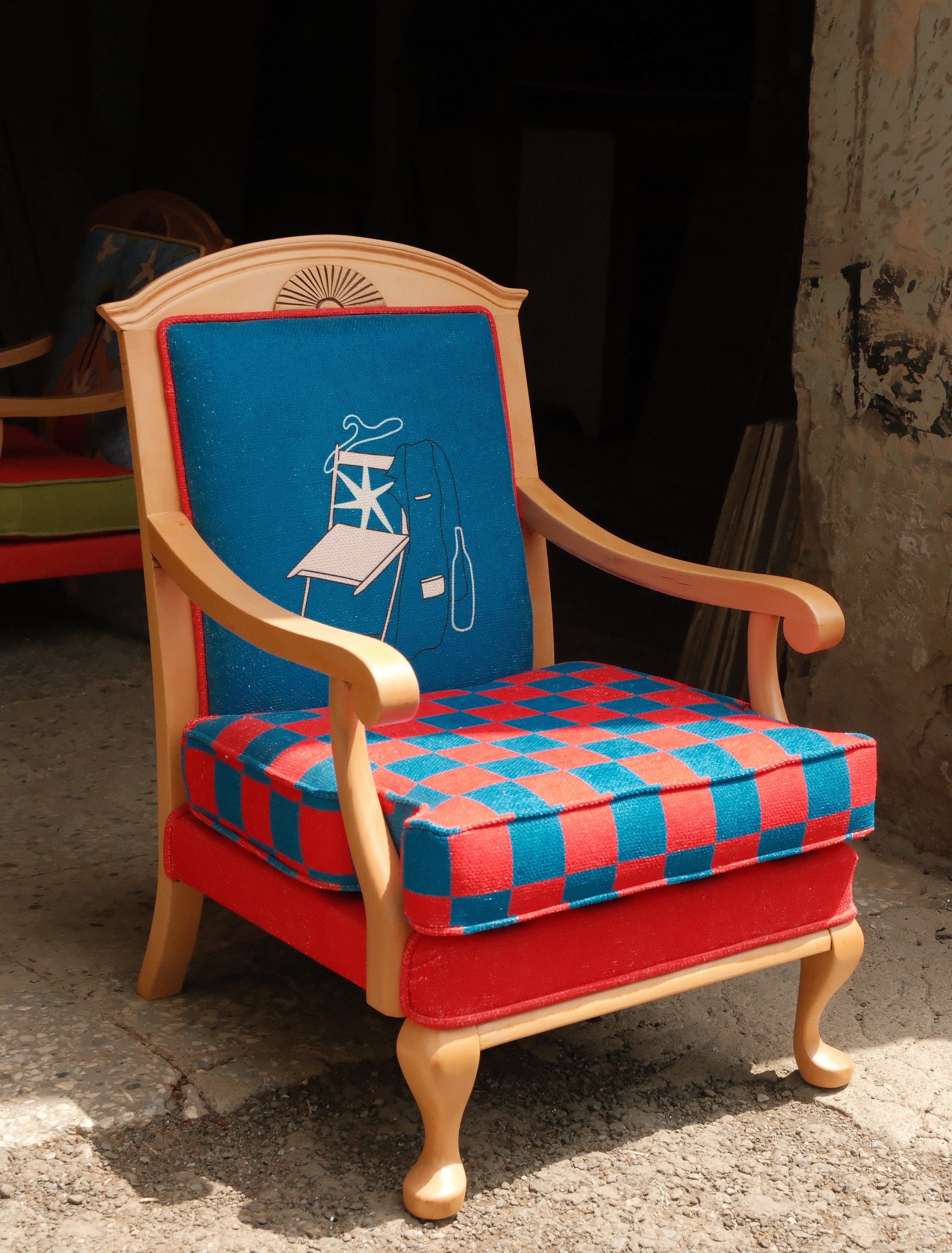 A vintage wooden armchair with a colorful patterned upholstery, featuring a red and blue checkered seat cushion, a solid red upholstered base, and a blue backrest with a white line-art illustration of a smoking figure.