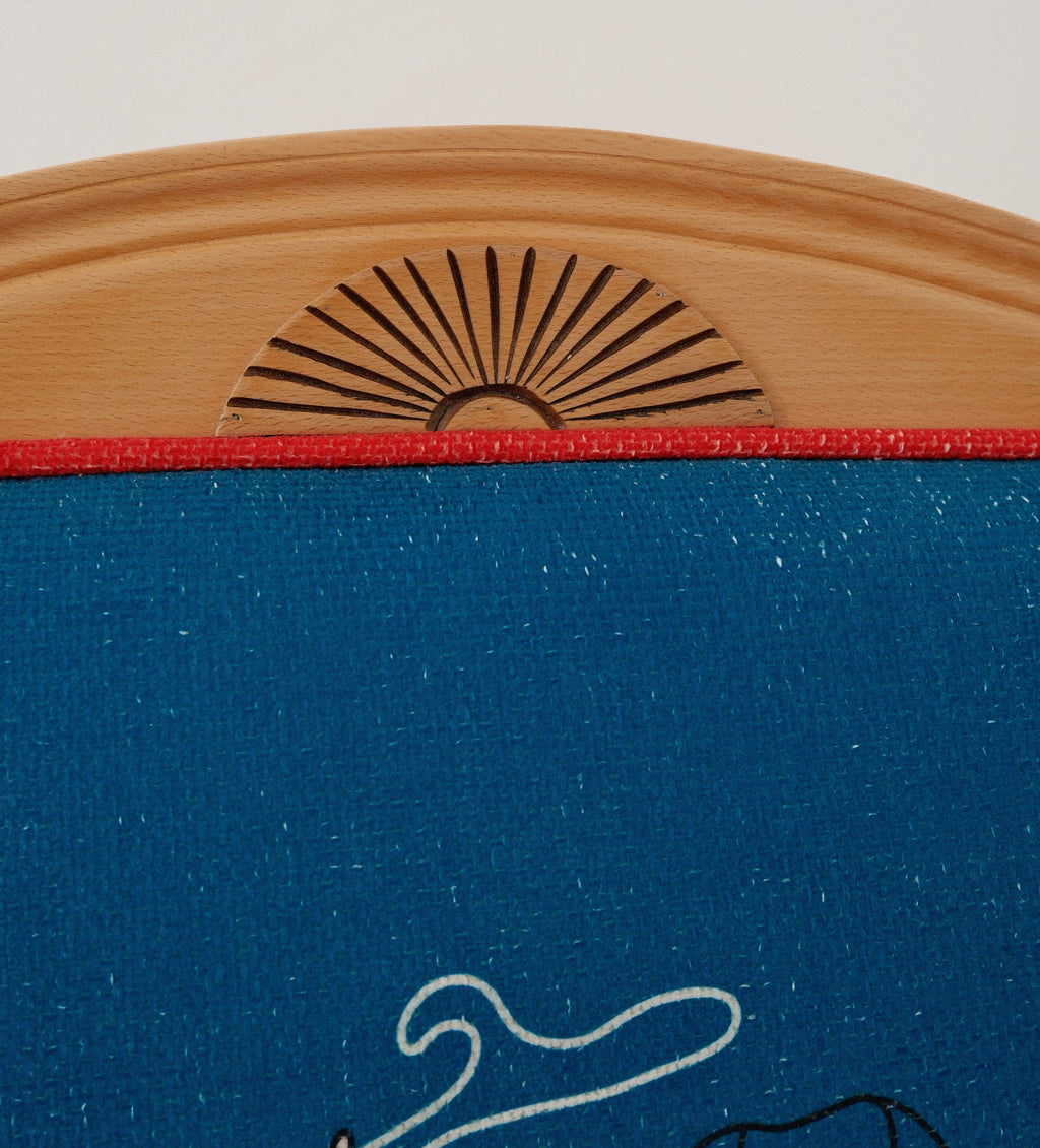 Close-up of a blue fabric-covered chair backrest with a natural wood top frame, featuring a laser-burned sun motif and a red horizontal trim line.