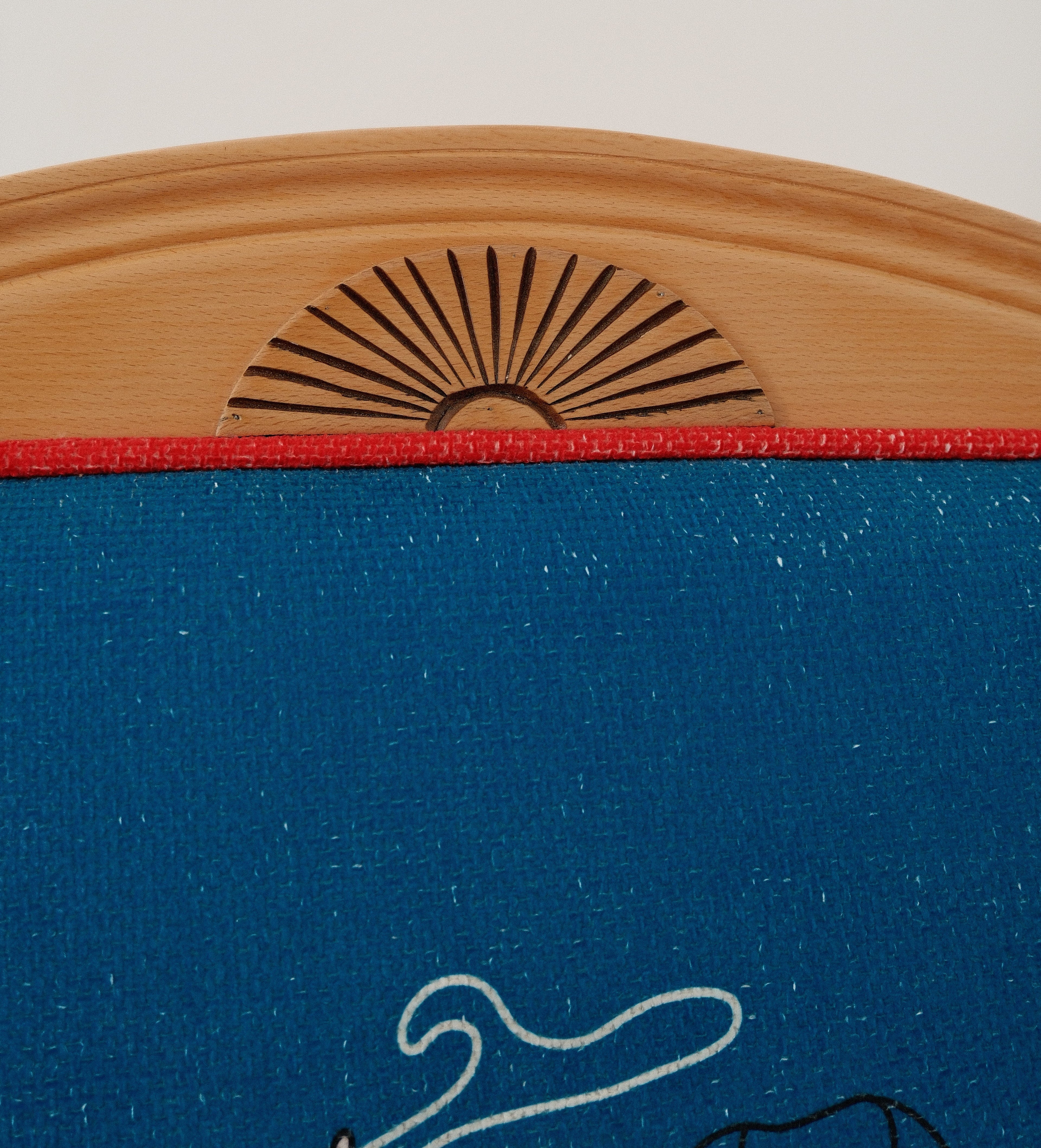 Close-up of a blue fabric-covered chair backrest with a natural wood top frame, featuring a laser-burned sun motif and a red horizontal trim line.