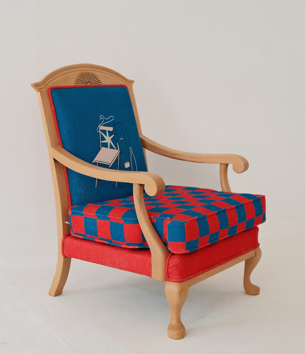 A vintage-style wooden armchair with a light natural wood frame, featuring a red and blue checkered seat cushion, a solid red upholstered base, and a blue upholstered backrest with a small white line-art illustration.