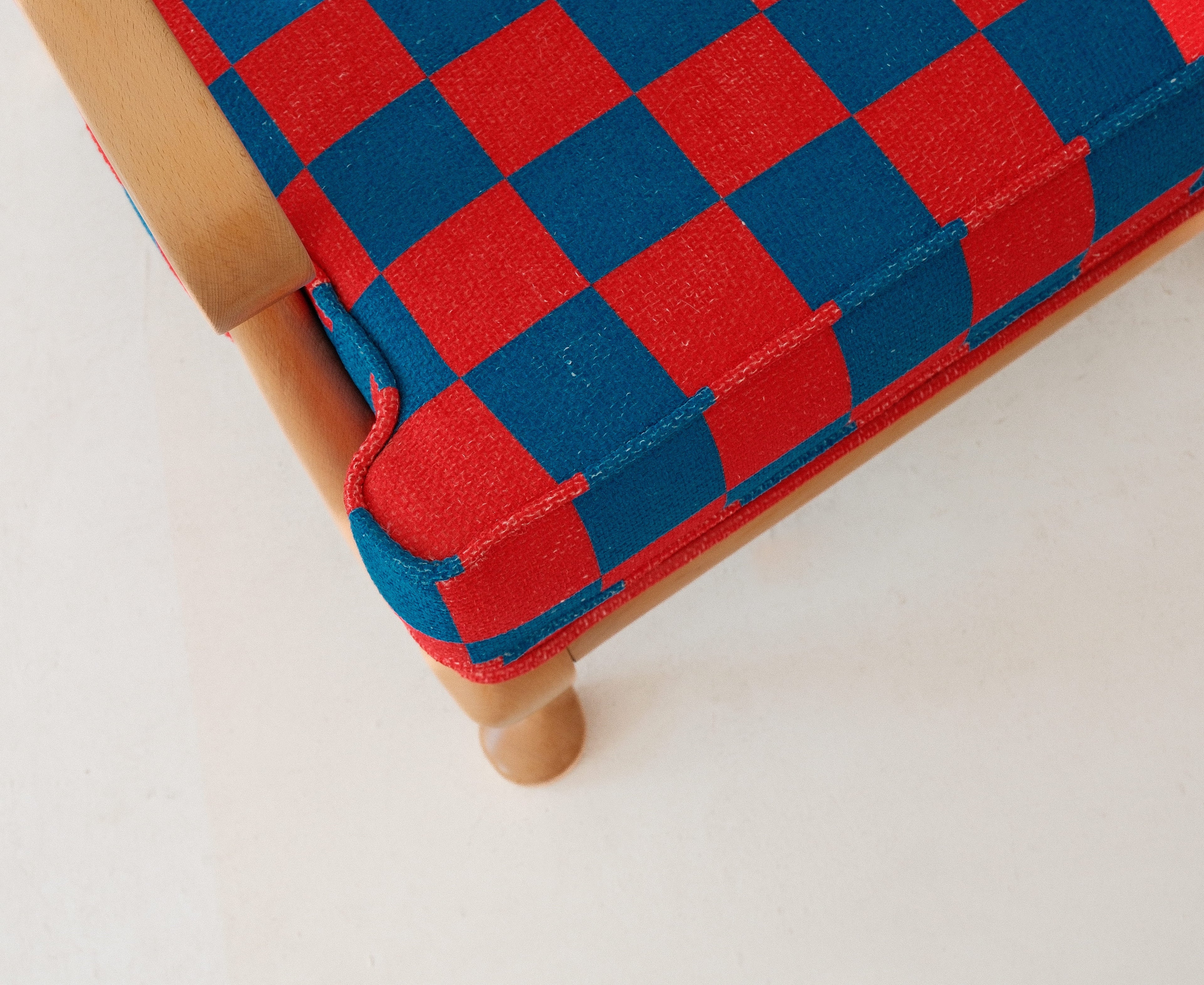 Close-up of an upholstered wooden chair corner showing a red and blue checkerboard fabric seat with a light natural wood frame on a white background.