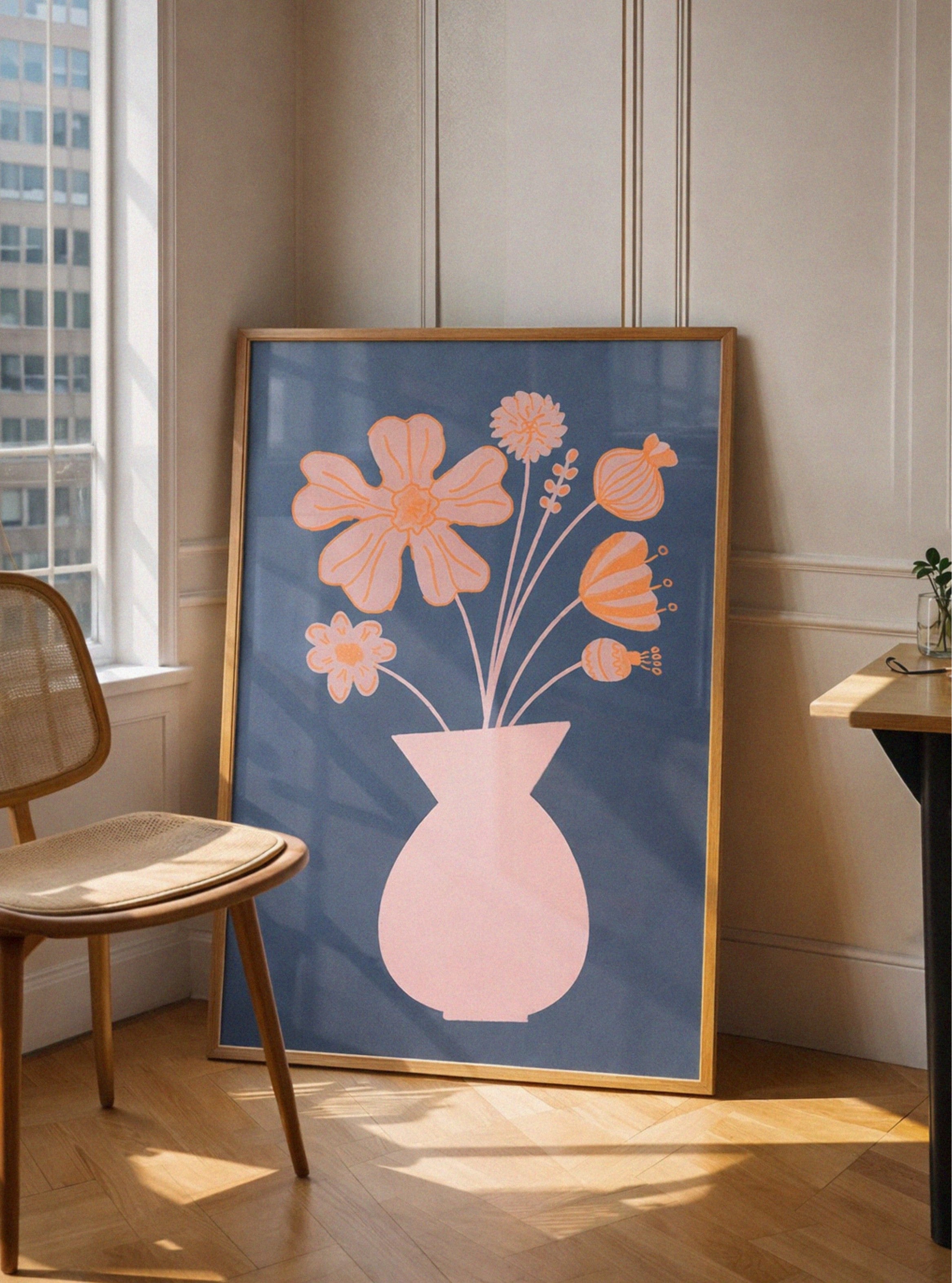 Framed floral artwork with a pink vase on a blue background in a room with a window and chair.