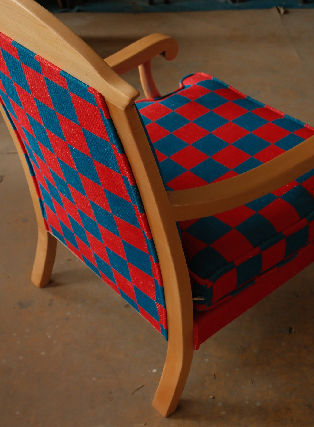 A wooden armchair with a red and blue diamond-patterned fabric upholstery, photographed in a workshop or studio setting on a concrete floor.