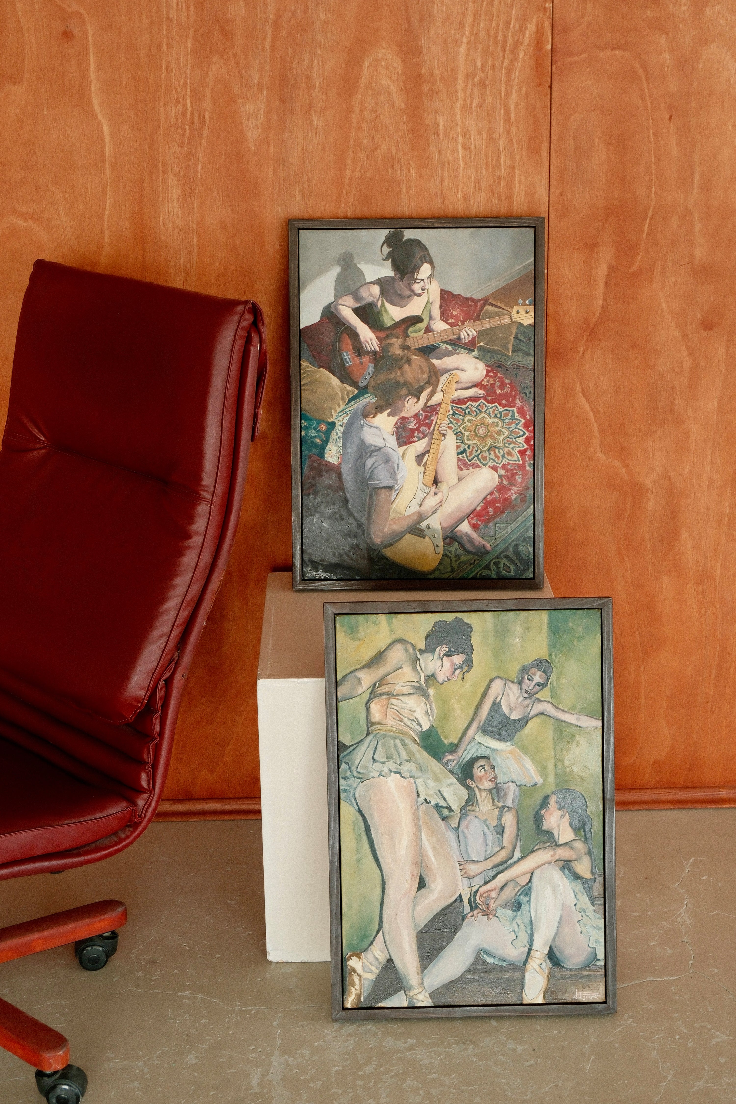 Two framed artworks on a stand against a wooden wall with a red chair.
