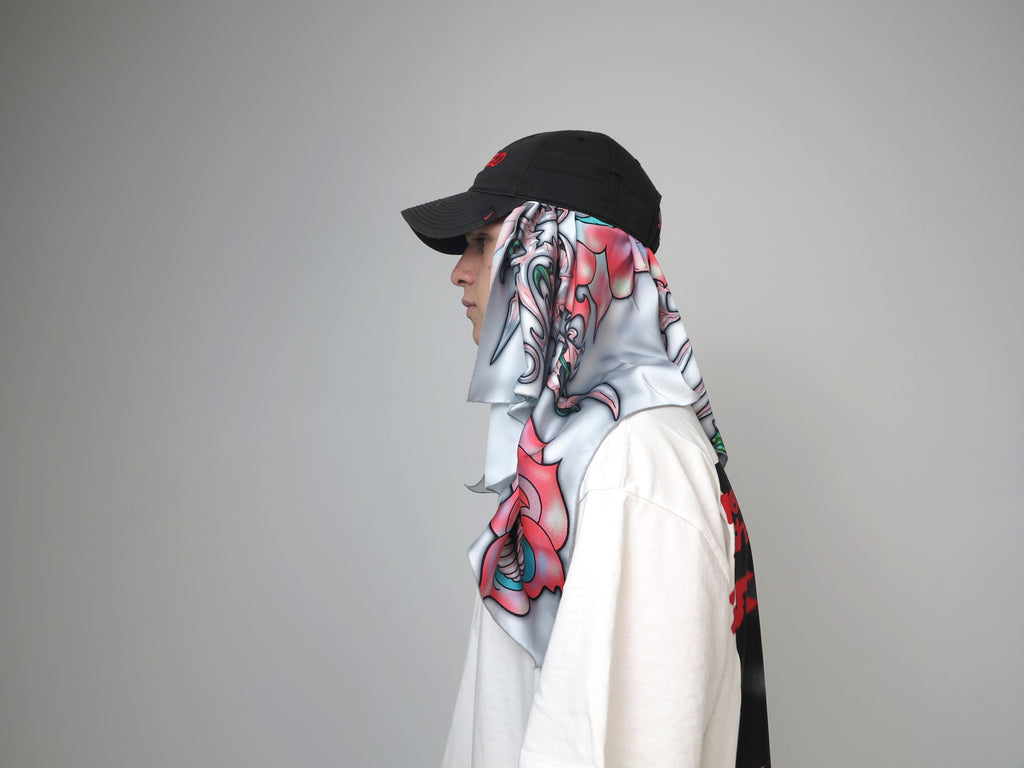 A person wearing a white shirt and a black cap, with a multicolored scarf featuring a floral pattern wrapped around the neck.