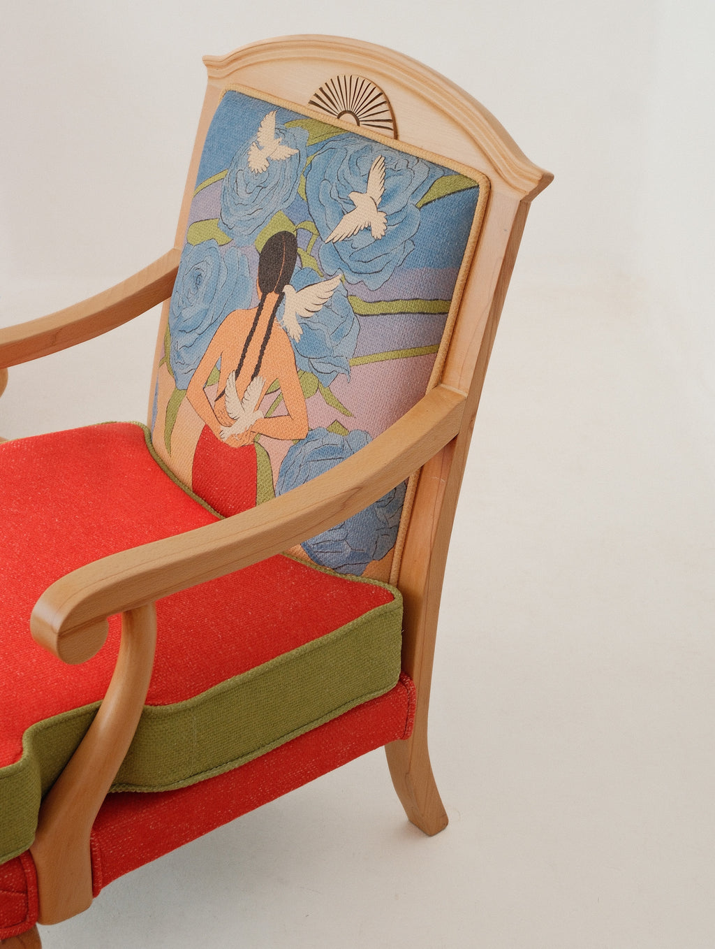 A close-up of an upholstered wooden armchair with a light natural wood frame, a red seat cushion, an olive green side bolster, and a highly detailed backrest panel featuring a woman in a red dress surrounded by blue flowers and white doves.