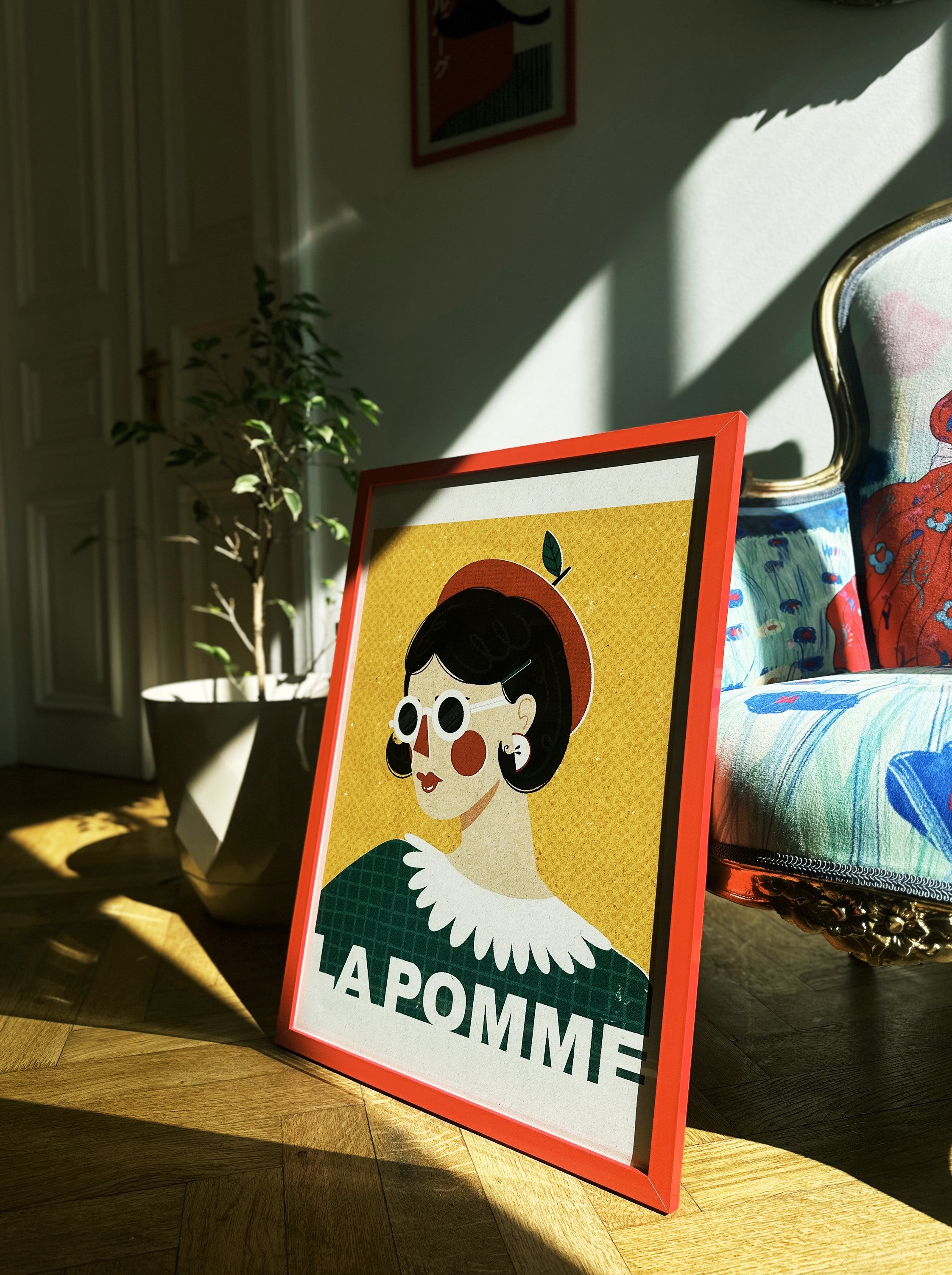 A framed poster with a colorful illustration, featuring a character with an apple accessory, displayed in a room with a window and a plant.