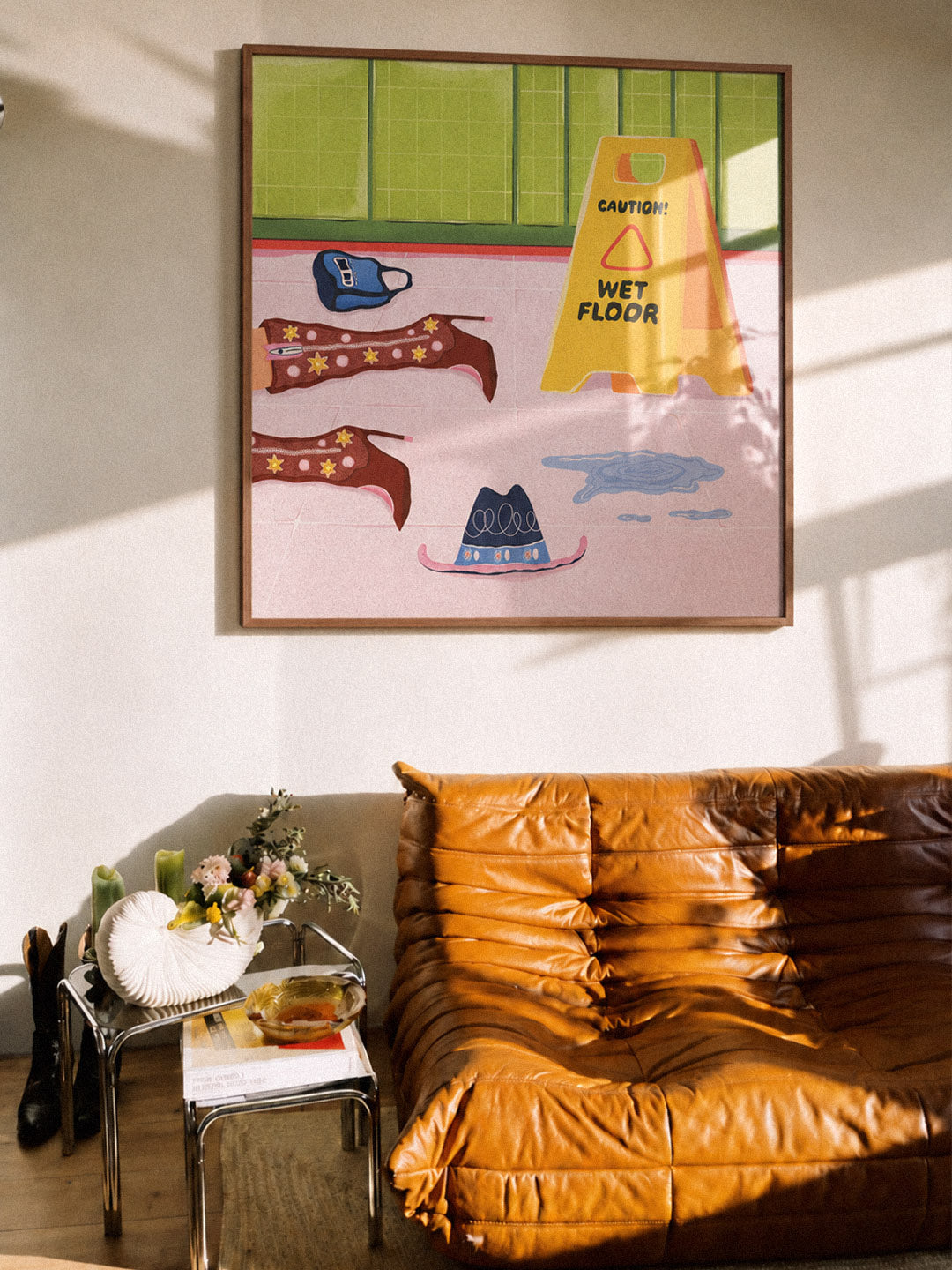 A framed art print featuring a whimsical wet floor scene with a yellow caution cone, blue shoes, and brown star-patterned ribbons, displayed on a wall above a tan leather sofa in a sunlit room. Poster made by Georgian company DAE