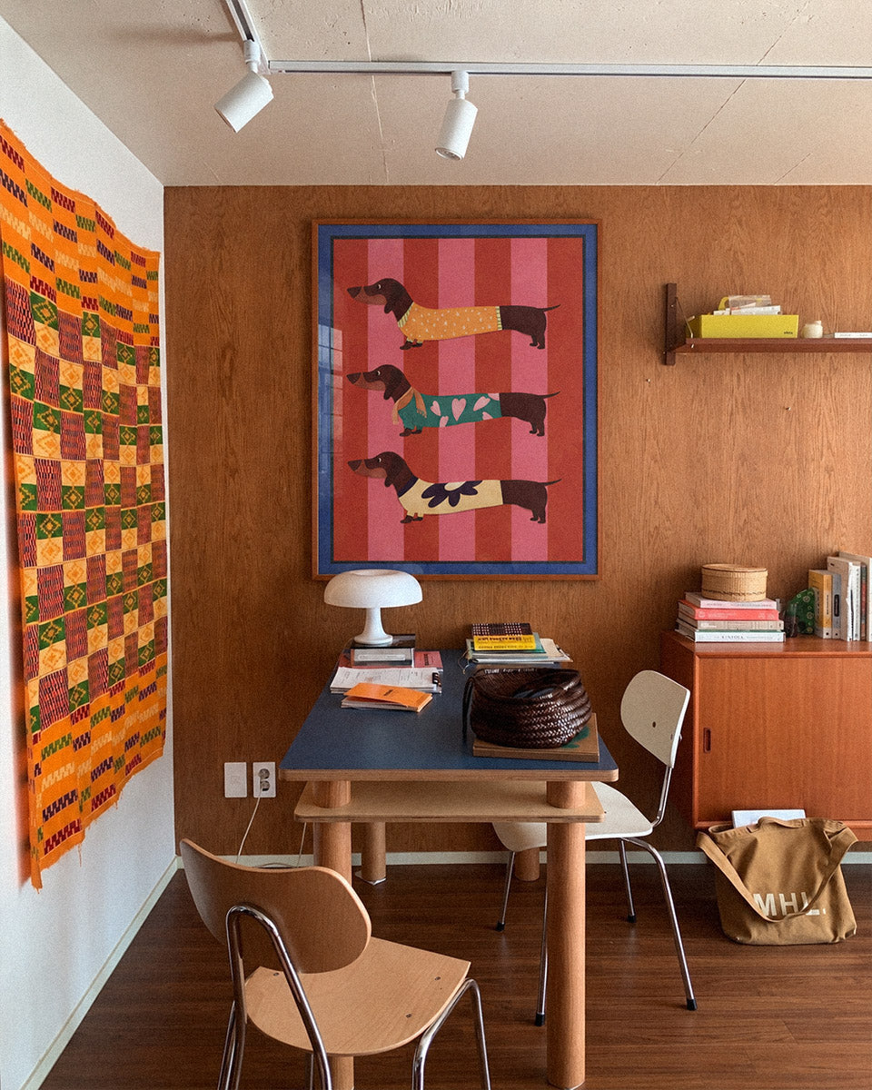 A framed art print featuring four dachshunds on a pink and red striped background, hung on a brown wood-paneled wall in a retro-styled room with a desk, chairs, and a colorful quilt.