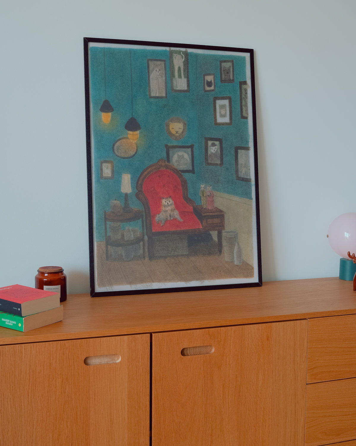 A framed art print depicting a cozy room scene with a red armchair, blue walls, and framed pictures, displayed on a wooden cabinet against a light wall.