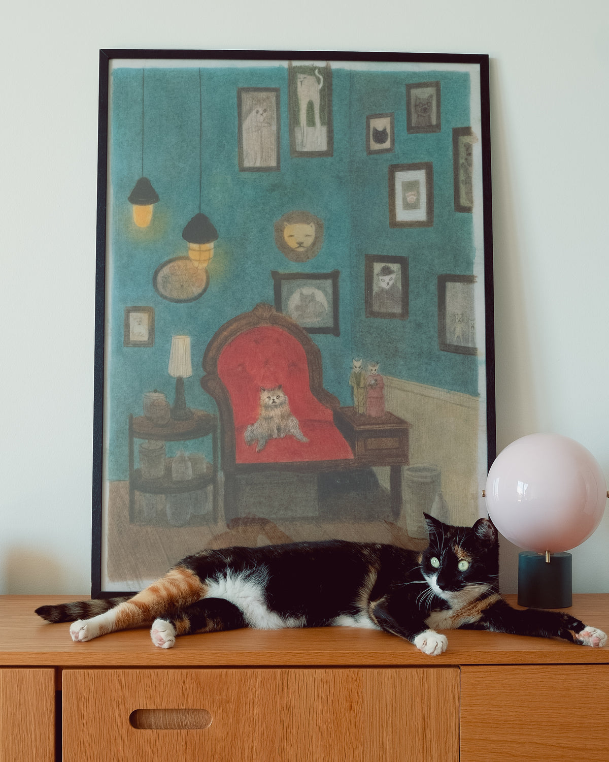 A framed art print depicting a cozy room with a cat in a red armchair, styled on a wooden cabinet with a black-and-white cat lying in front of it and a pink globe lamp on the right.