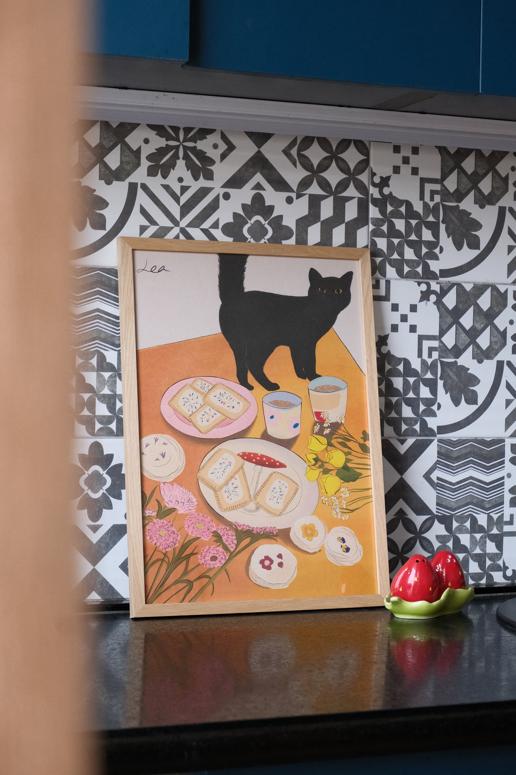 Illustrated poster with a cat and cookies on an orange background, placed on a patterned surface.
