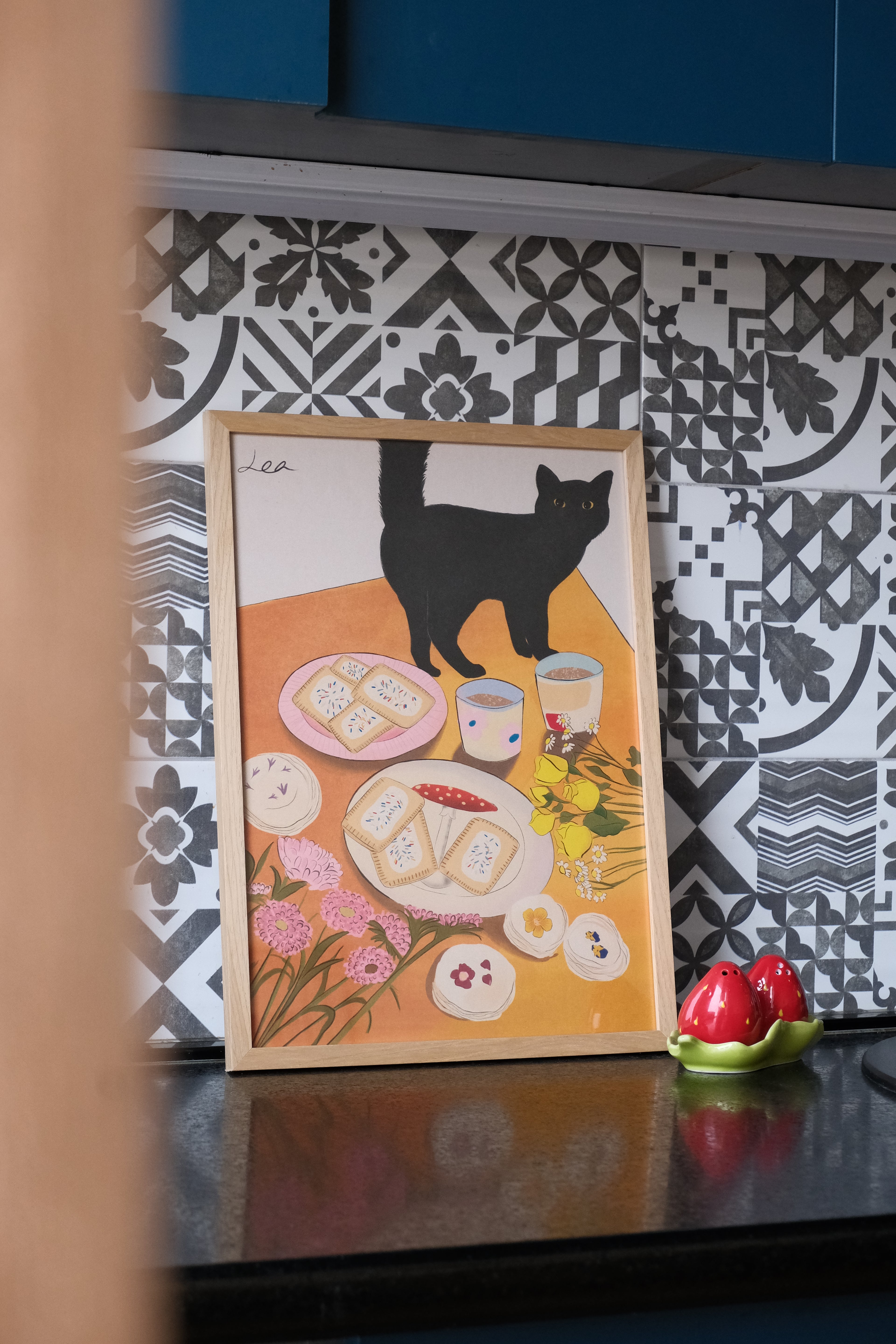Illustrated poster with a cat and cookies on an orange background, placed on a patterned surface.