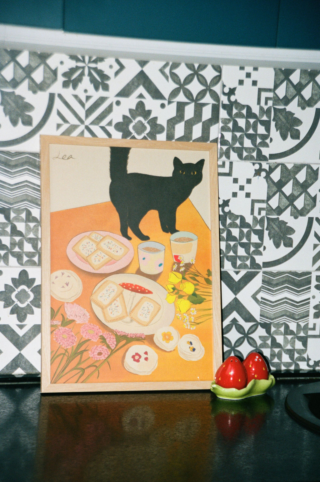 A poster featuring a black cat with a design by Lea Woo. The illustration includes plates of food, cups of tea, and floral elements in soft colors.