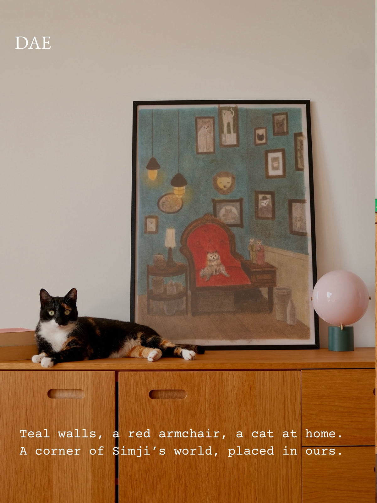 A framed art print depicting a cozy room corner with teal walls, a red armchair, and a cat, displayed on a wooden cabinet next to a black-and-white cat and a pink globe lamp. The text 'DAE' appears in the top left, and descriptive text is at the bottom.