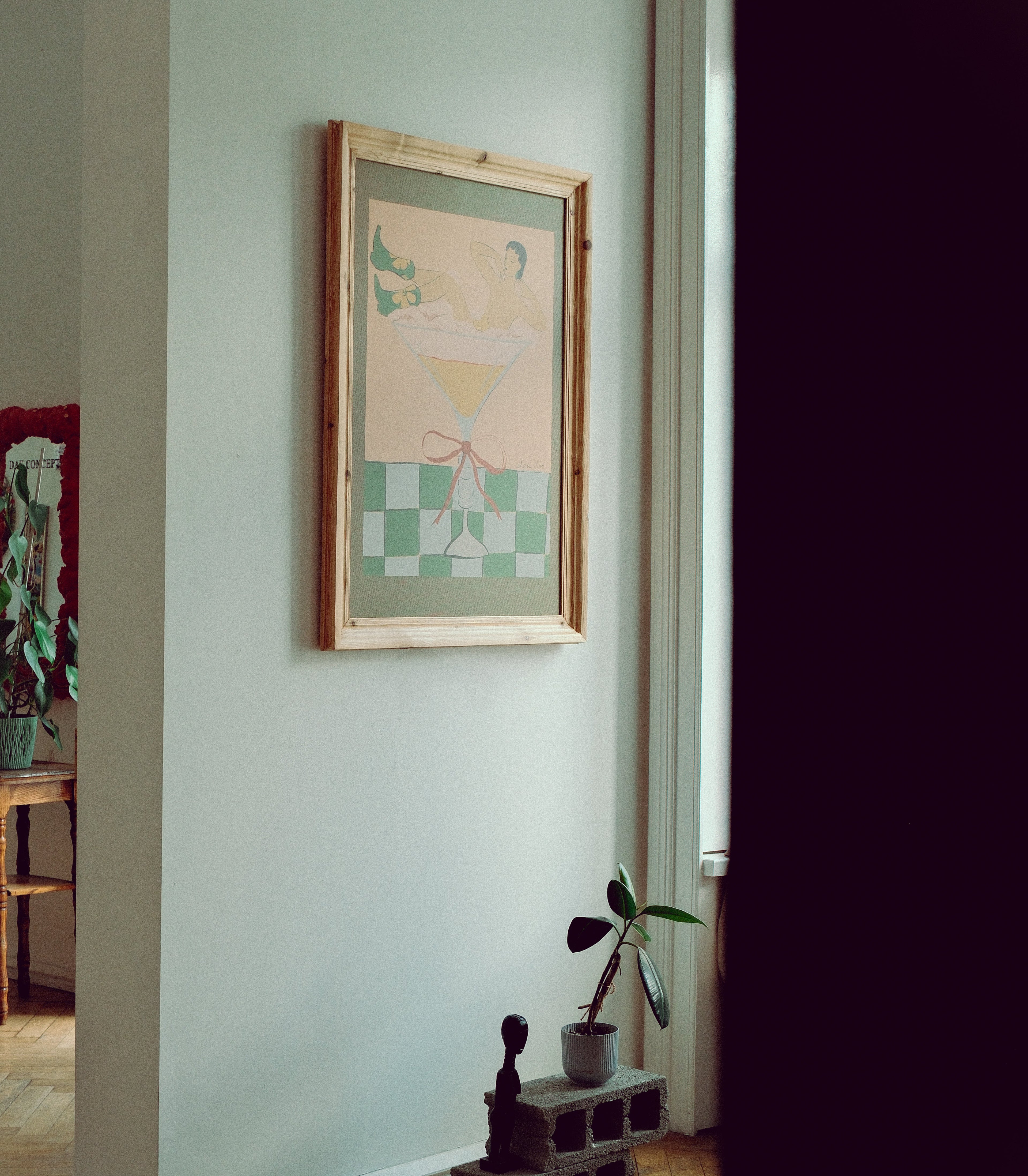 A framed art print featuring a stylized illustration of a woman in a bathtub, hung on a light gray wall in a home interior next to a small potted plant.