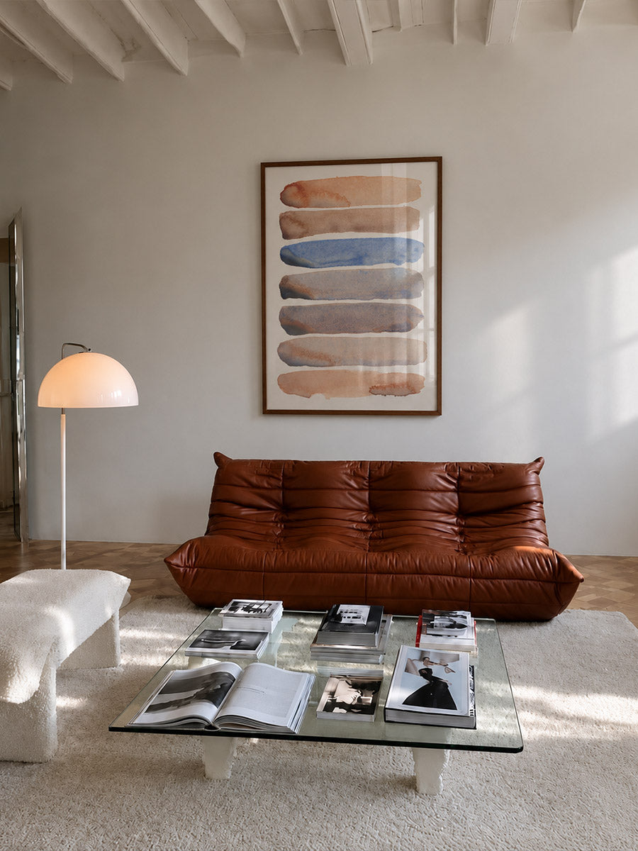 A modern living room featuring a brown leather chaise lounge sofa, a glass coffee table with magazines, a white ottoman, and a framed abstract art print on a light gray wall.