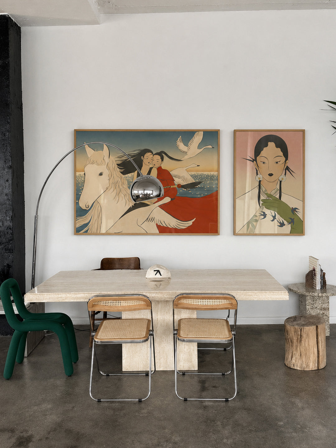 A styled dining room interior featuring two framed art prints on a white wall above a light wood table and woven-back chairs. The left print shows a white horse and rider with a red cloak, and the right print shows an illustrated woman with a green bird.