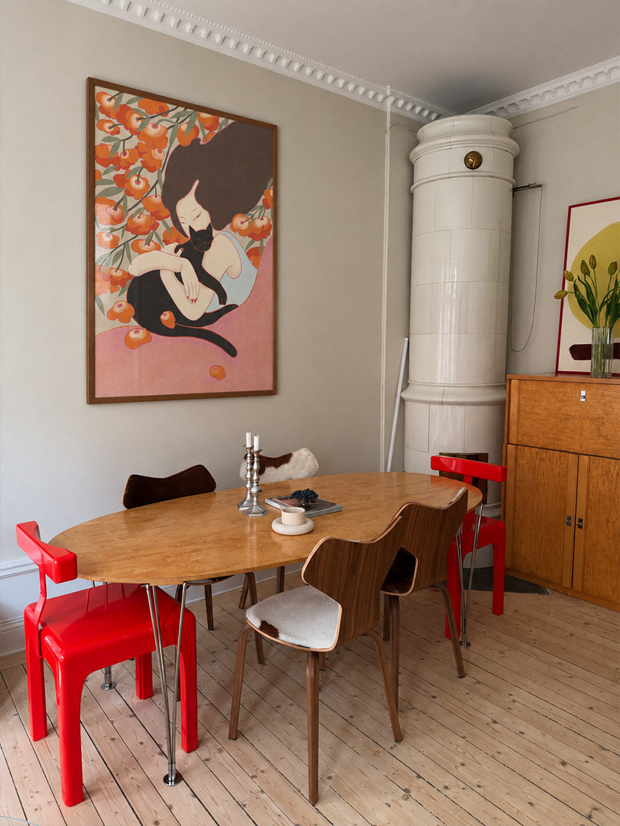 A styled dining room corner featuring a light wood oval table surrounded by red and wooden chairs, with a large framed art print on the wall depicting a woman among flowers, poster by DAE x Leah.